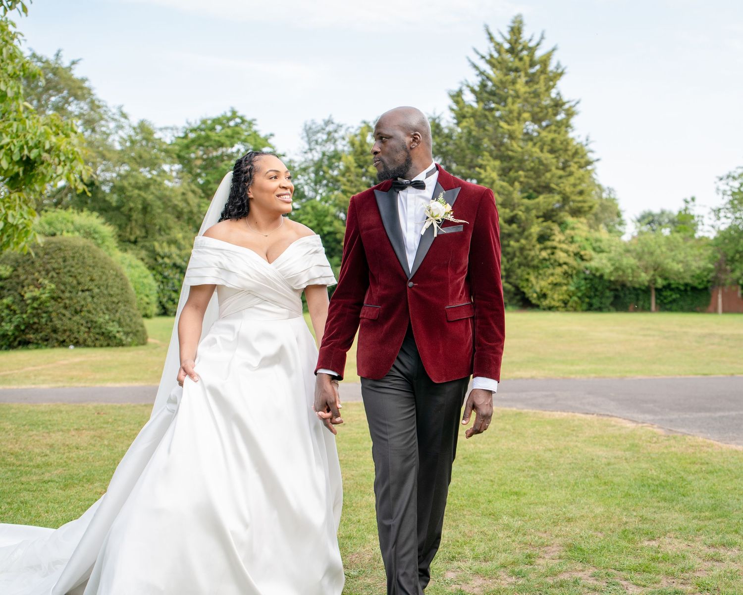 Happy newlyweds walk hand in hand outdoors, bride in white gown, groom in red velvet blazer and bow tie.