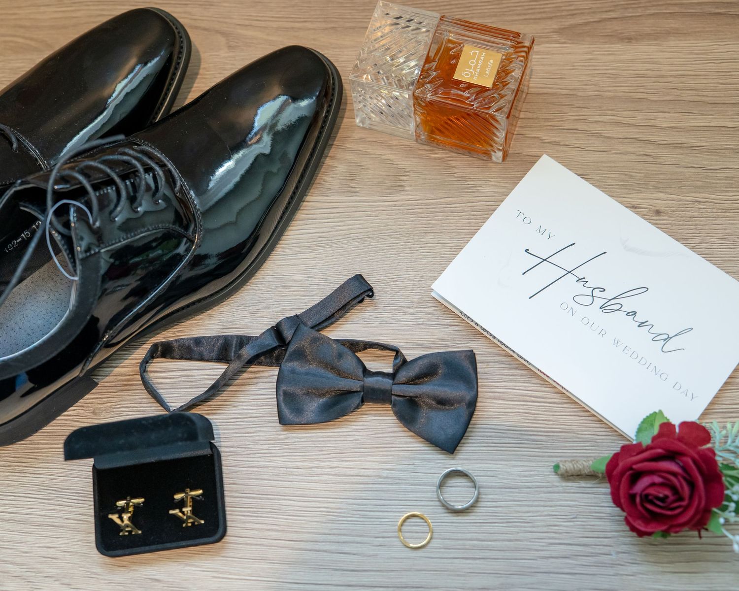Groom wedding flat lay with black patent shoes, bow tie, cufflinks, rings, cologne, red rose, and husband card.