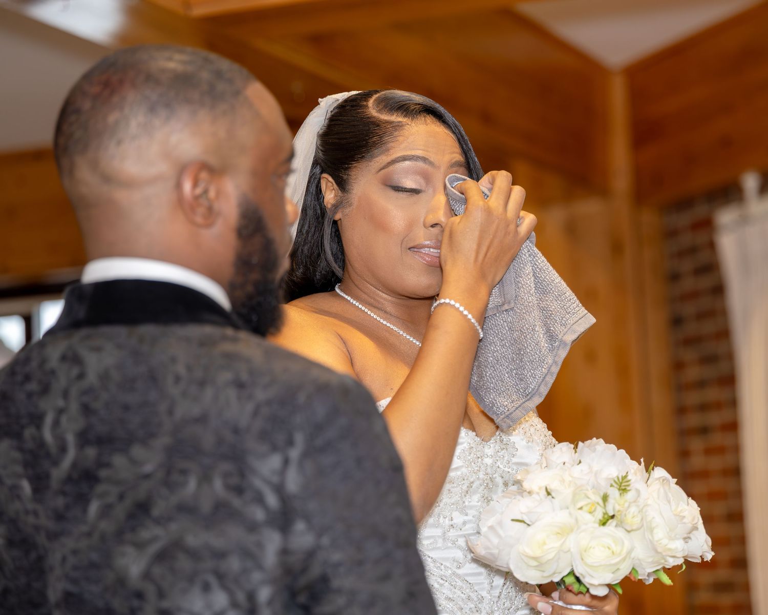 An emotional bride in a strapless lace gown dabs tears with a handkerchief while holding white roses during the ceremony.