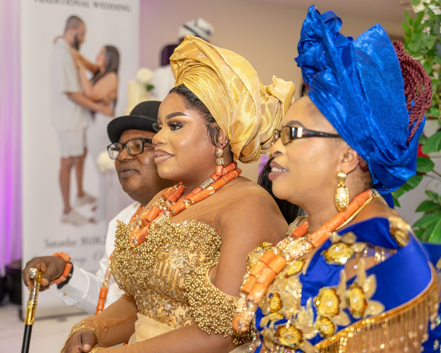 Three guests in vibrant Nigerian traditional wedding attire, featuring gold gele headwrap and blue royal aso-oke, smiling at ceremony.