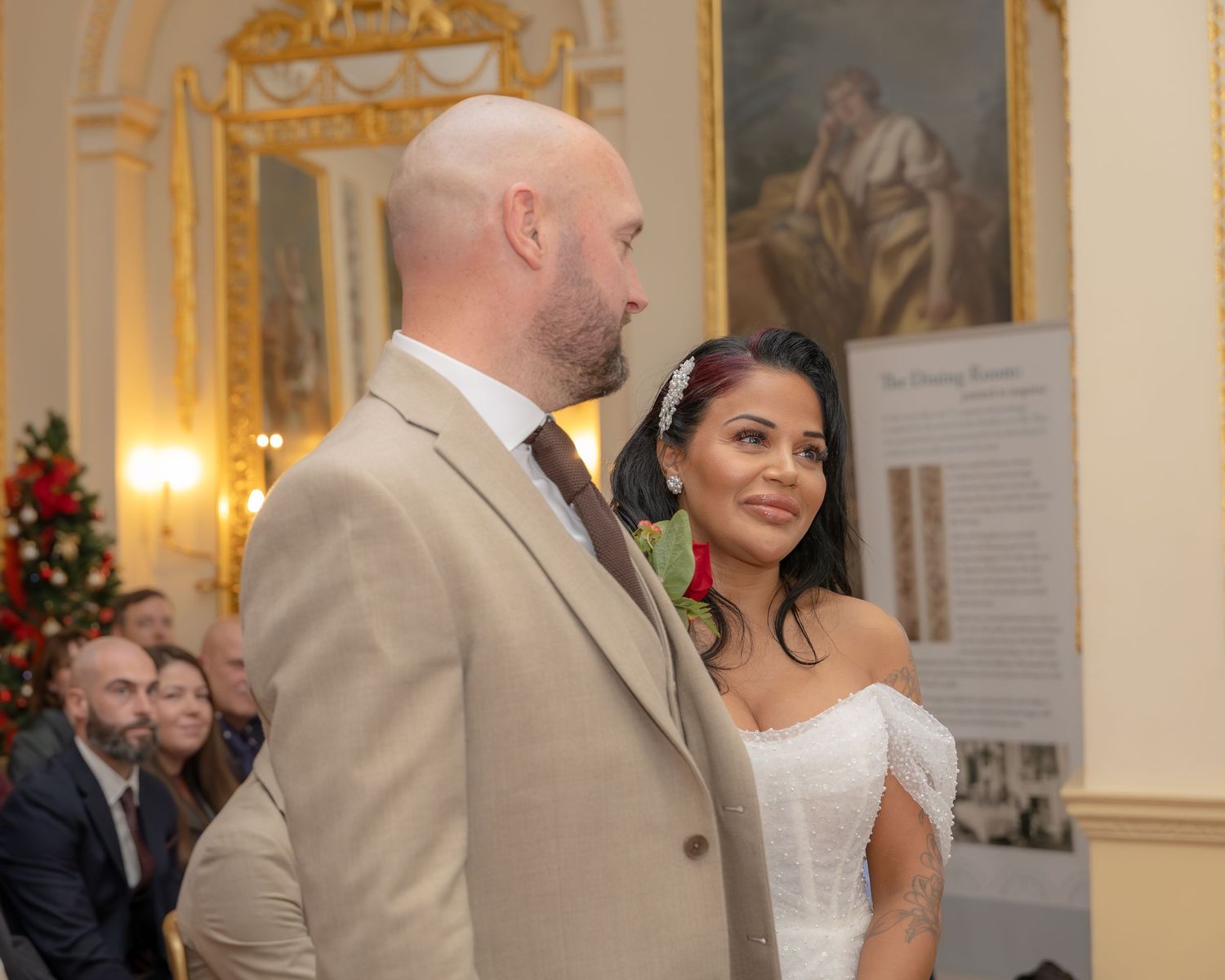 Couple at wedding ceremony with ornate interior, groom in tan suit and bride in white off-shoulder dress holding hands.