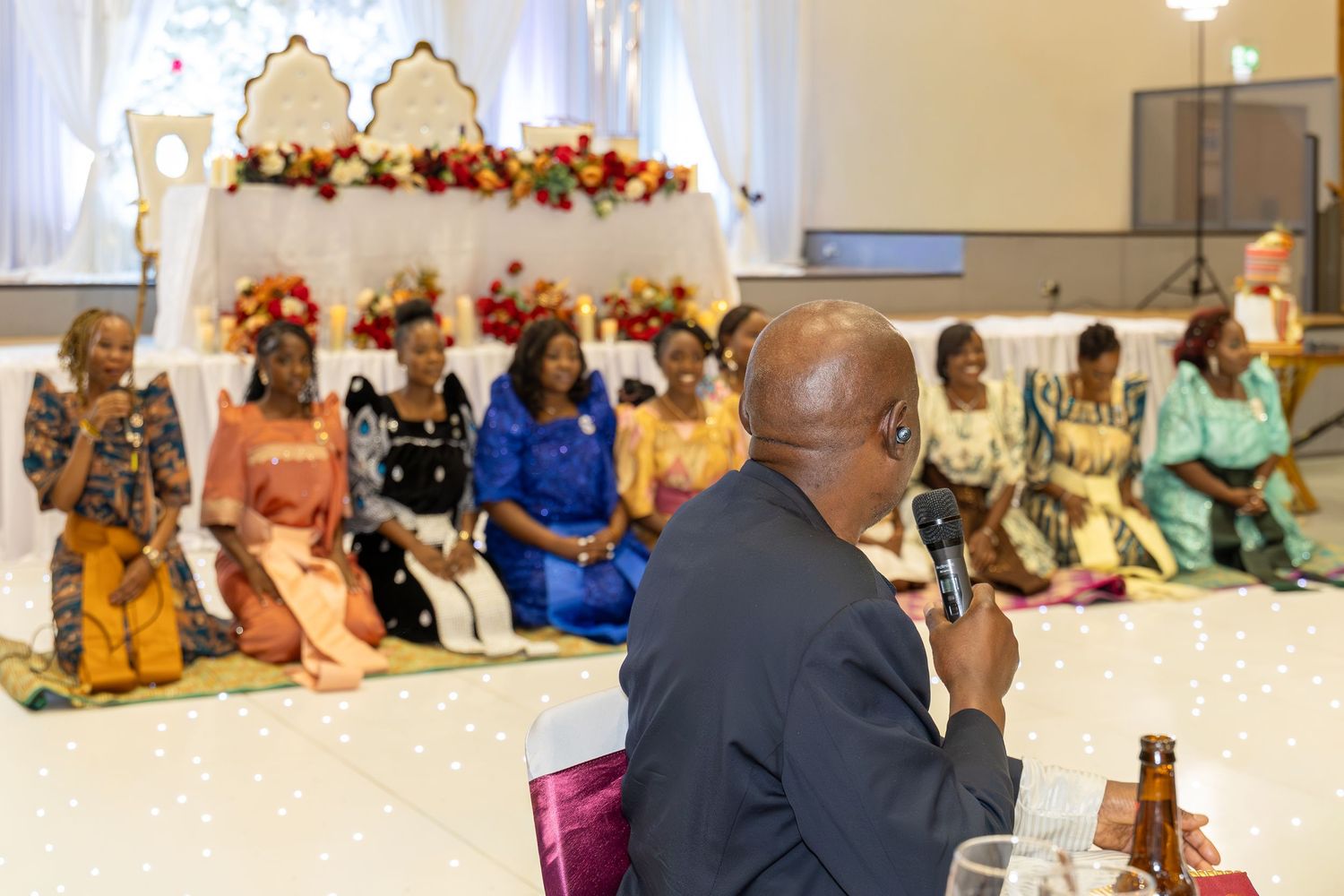 Man speaks into microphone at African cultural ceremony while women in traditional attire sit on floor before floral backdrop.