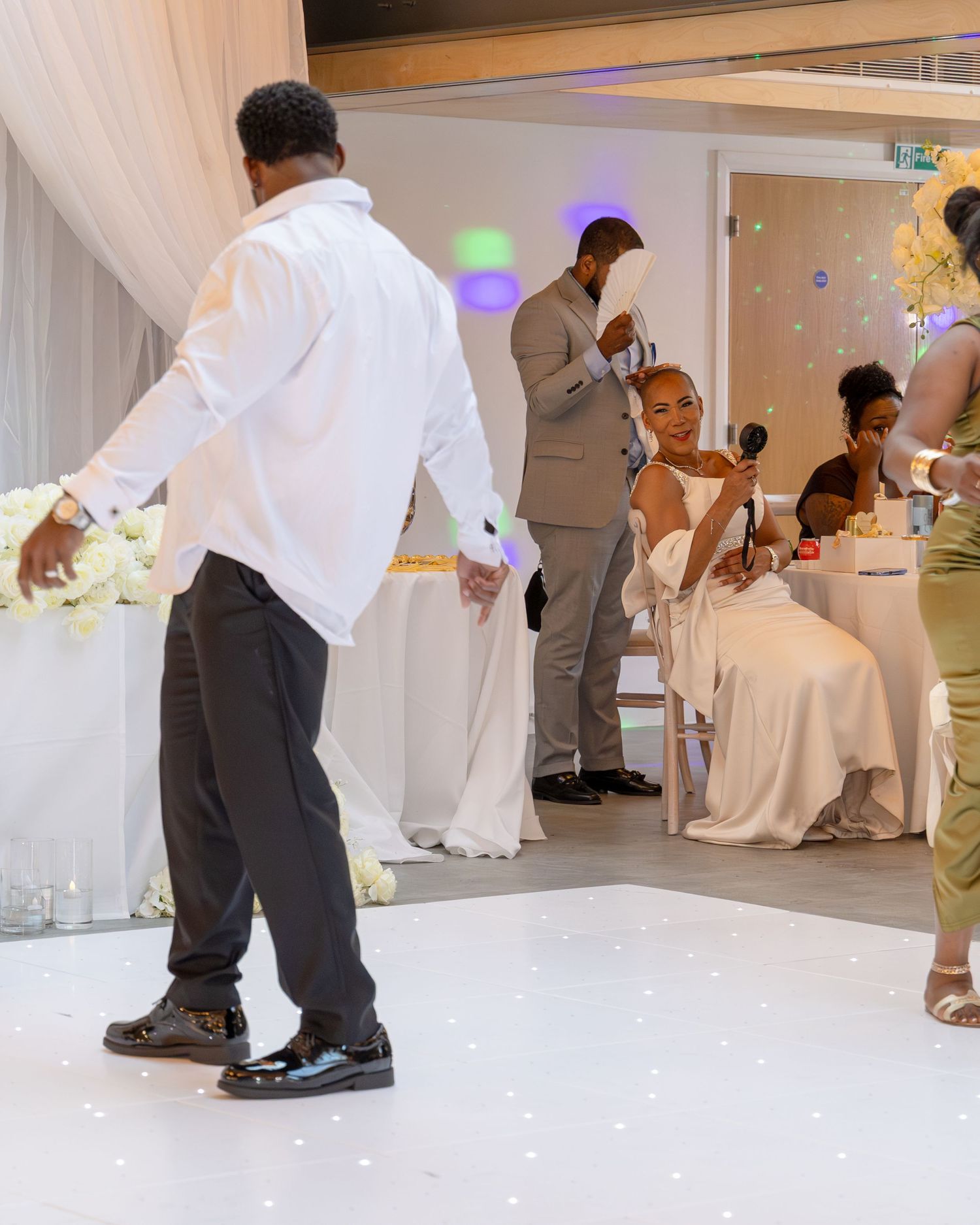 Bride in white gown seated at wedding reception as groom dances on white dance floor in elegant venue.
