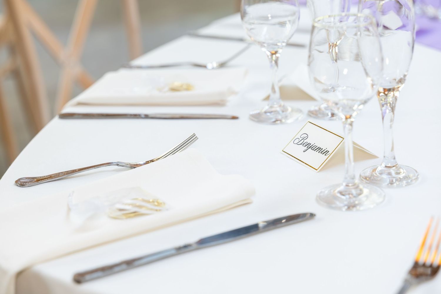 Elegant wedding table setting with white linen, crystal glasses, silverware, and a name card on a napkin.