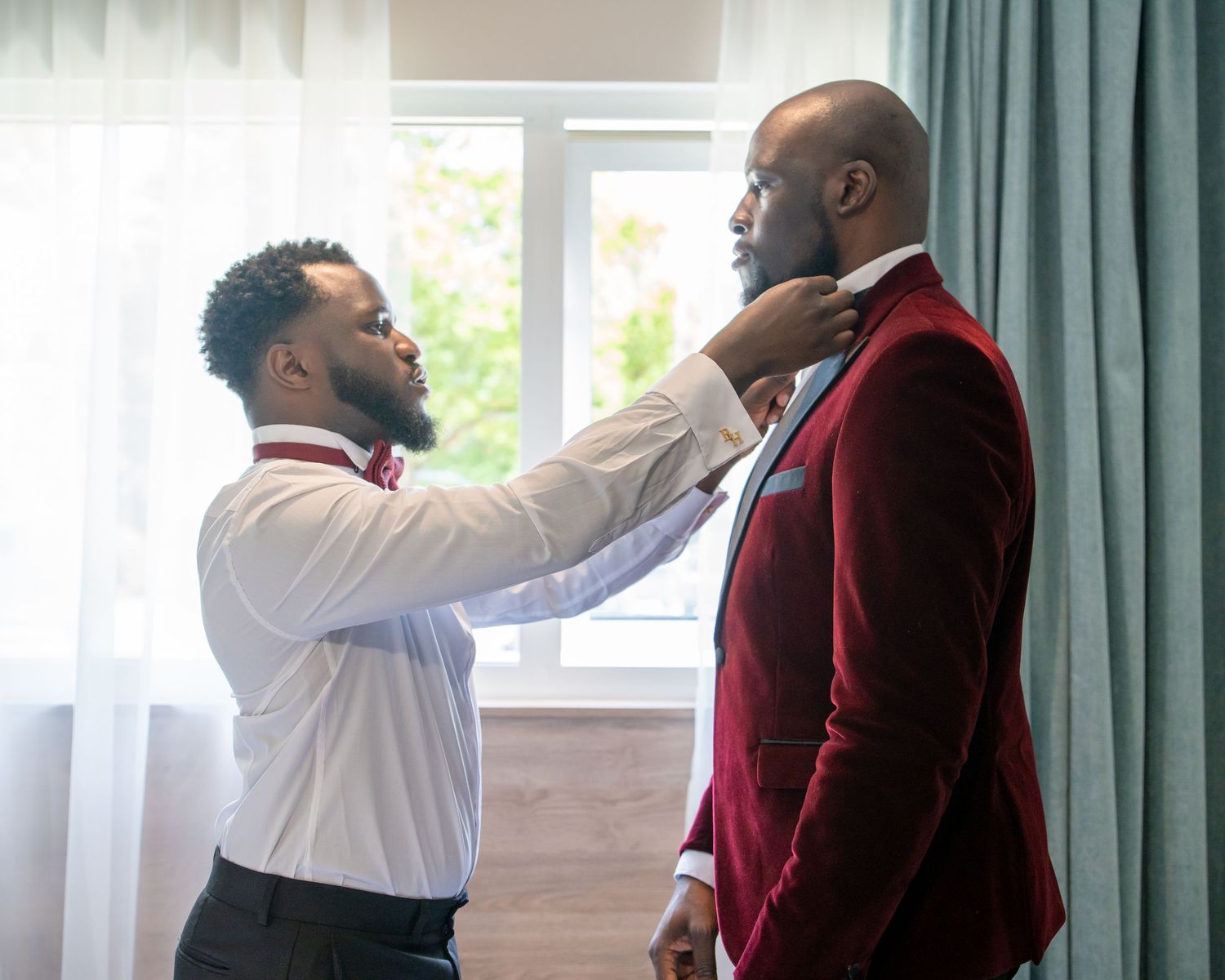Two well-dressed Black men adjusting a tie and collar before a formal wedding ceremony indoors.