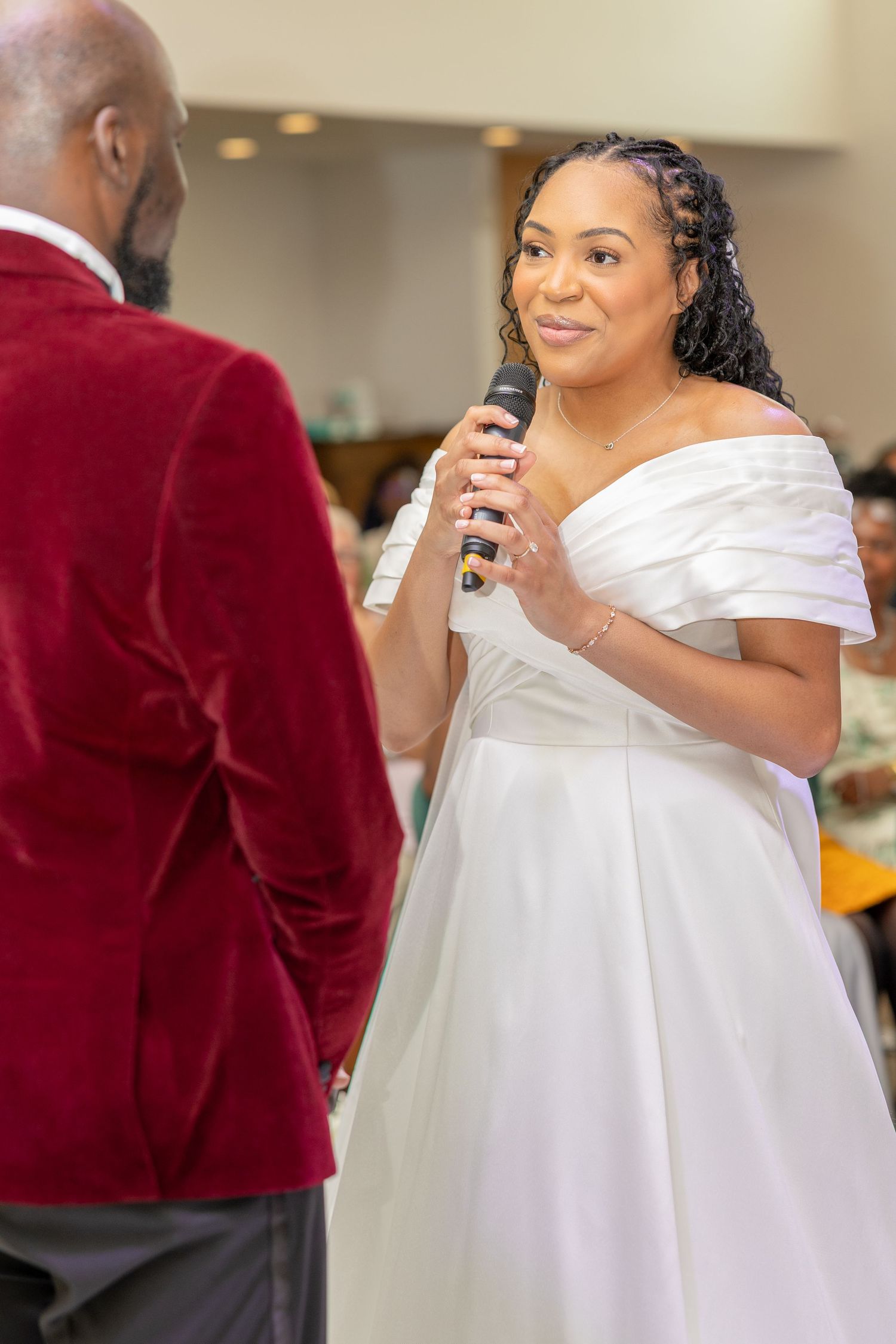 Bride in white off-shoulder gown speaks into microphone at wedding reception, facing groom in red velvet blazer.