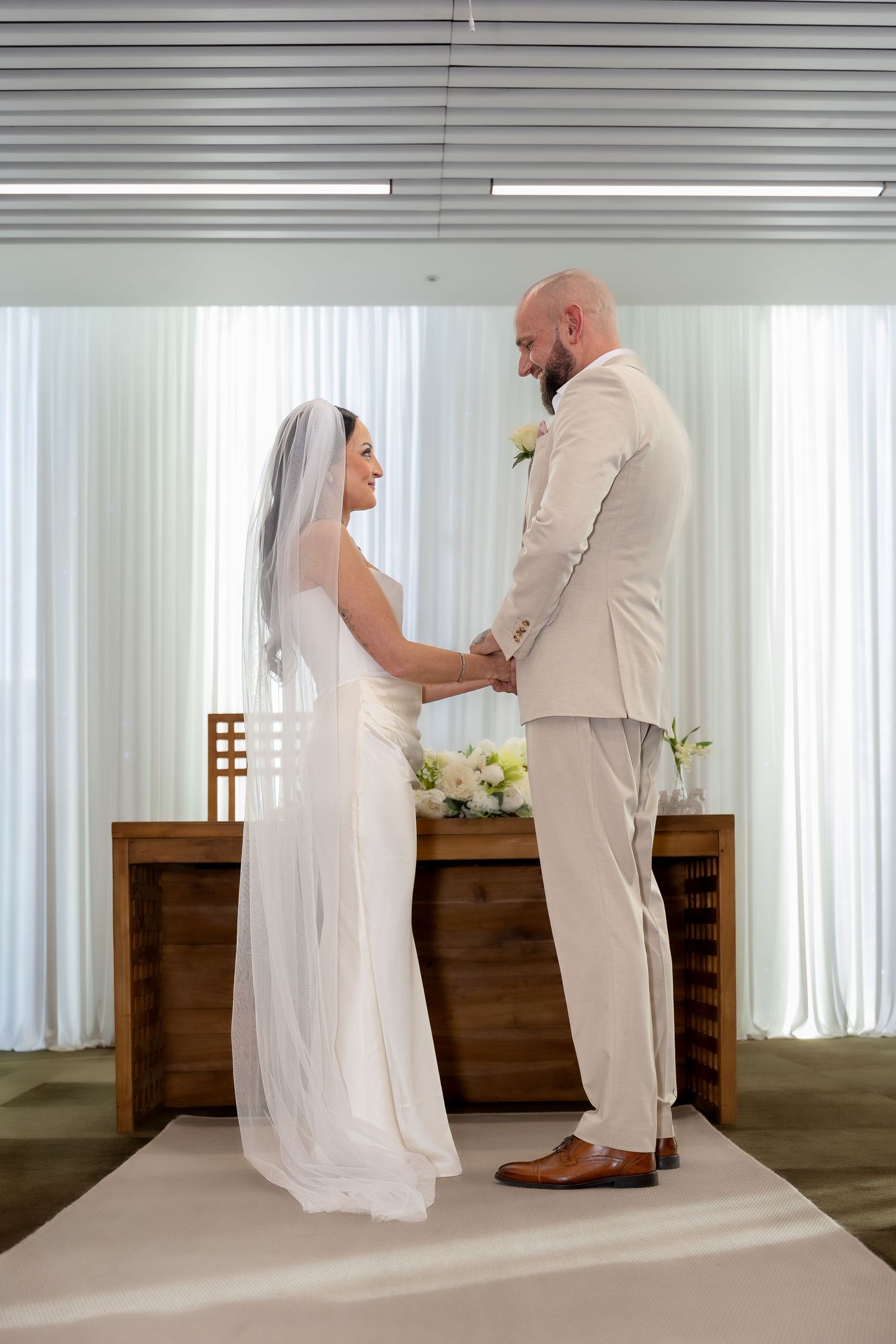Bride and groom hold hands facing each other at the altar in a bright modern wedding ceremony venue.