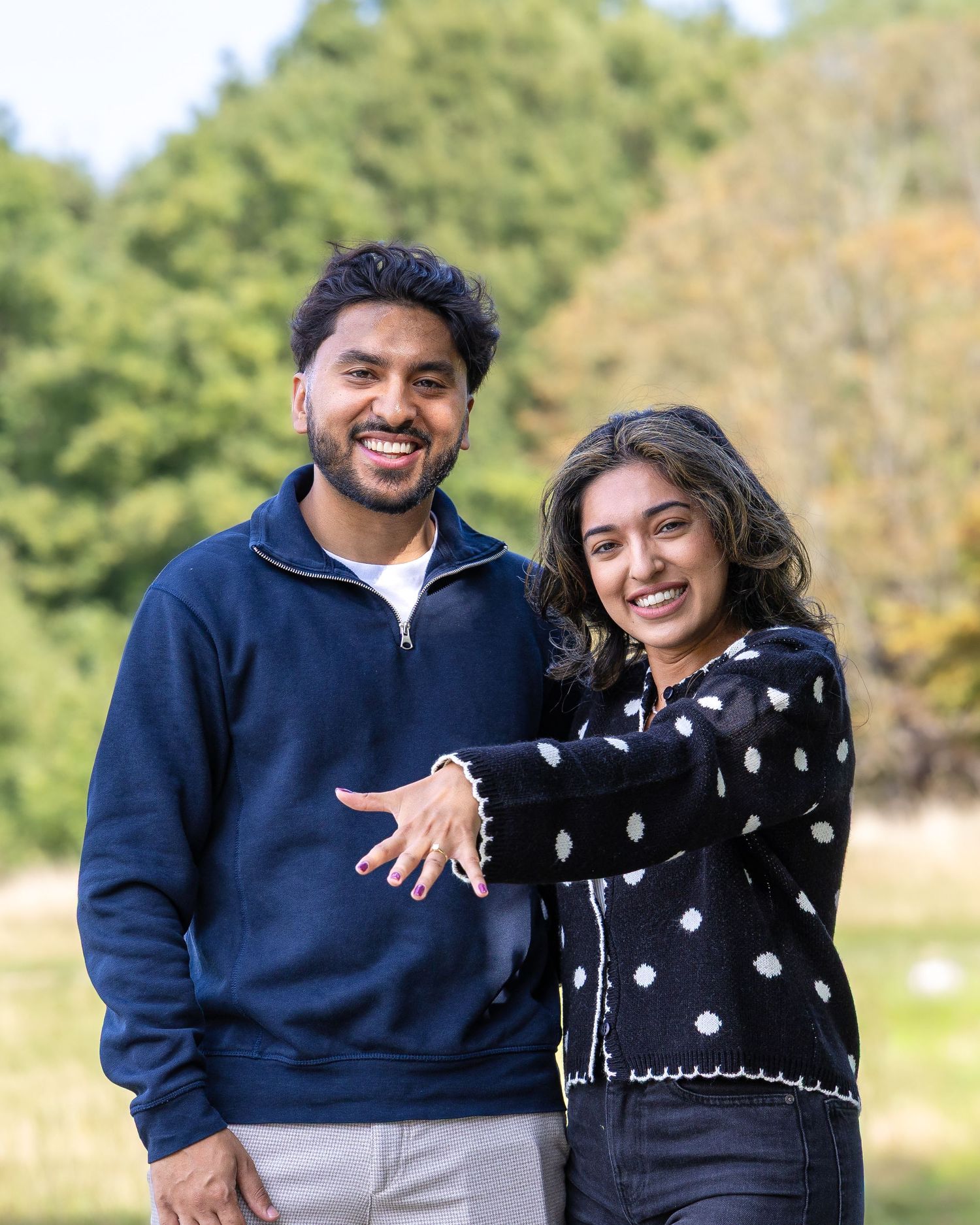 Smiling engaged couple posing outdoors showing off engagement ring in a green park setting.