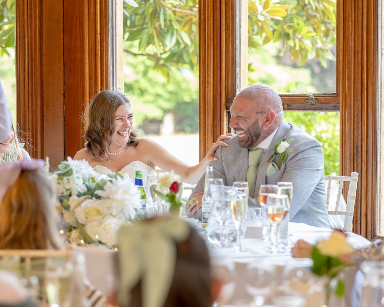 Bride and groom laughing joyfully at wedding reception table adorned with white floral centerpieces in elegant wood-paneled venue.