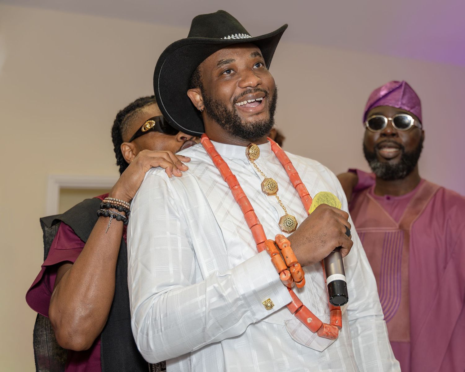 Smiling groom in black cowboy hat and coral beaded necklace enjoys Nigerian traditional wedding celebration.