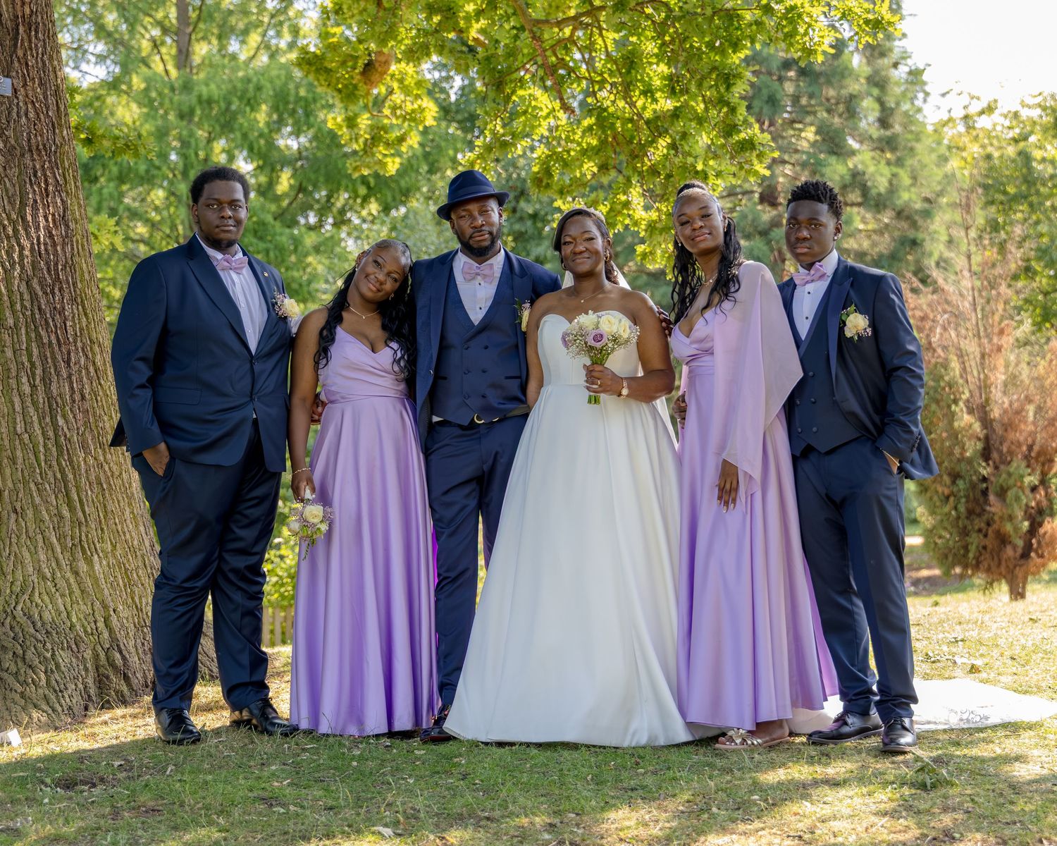 Wedding party poses outdoors under a large tree, bride in white gown flanked by groomsmen in navy suits and bridesmaids in lavender dresses.