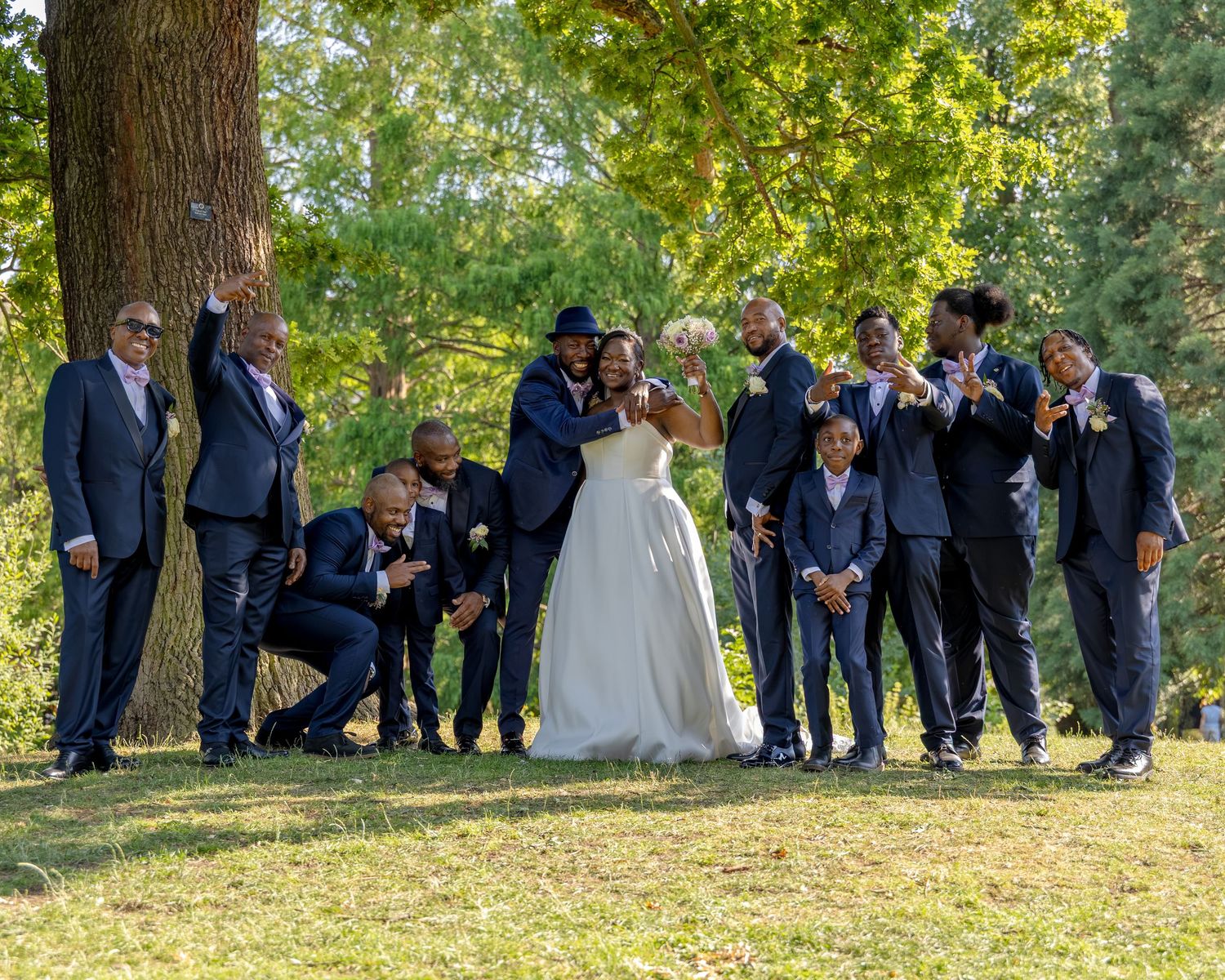 Wedding party in navy suits poses outdoors under a large tree with bride in white gown on a sunny day.