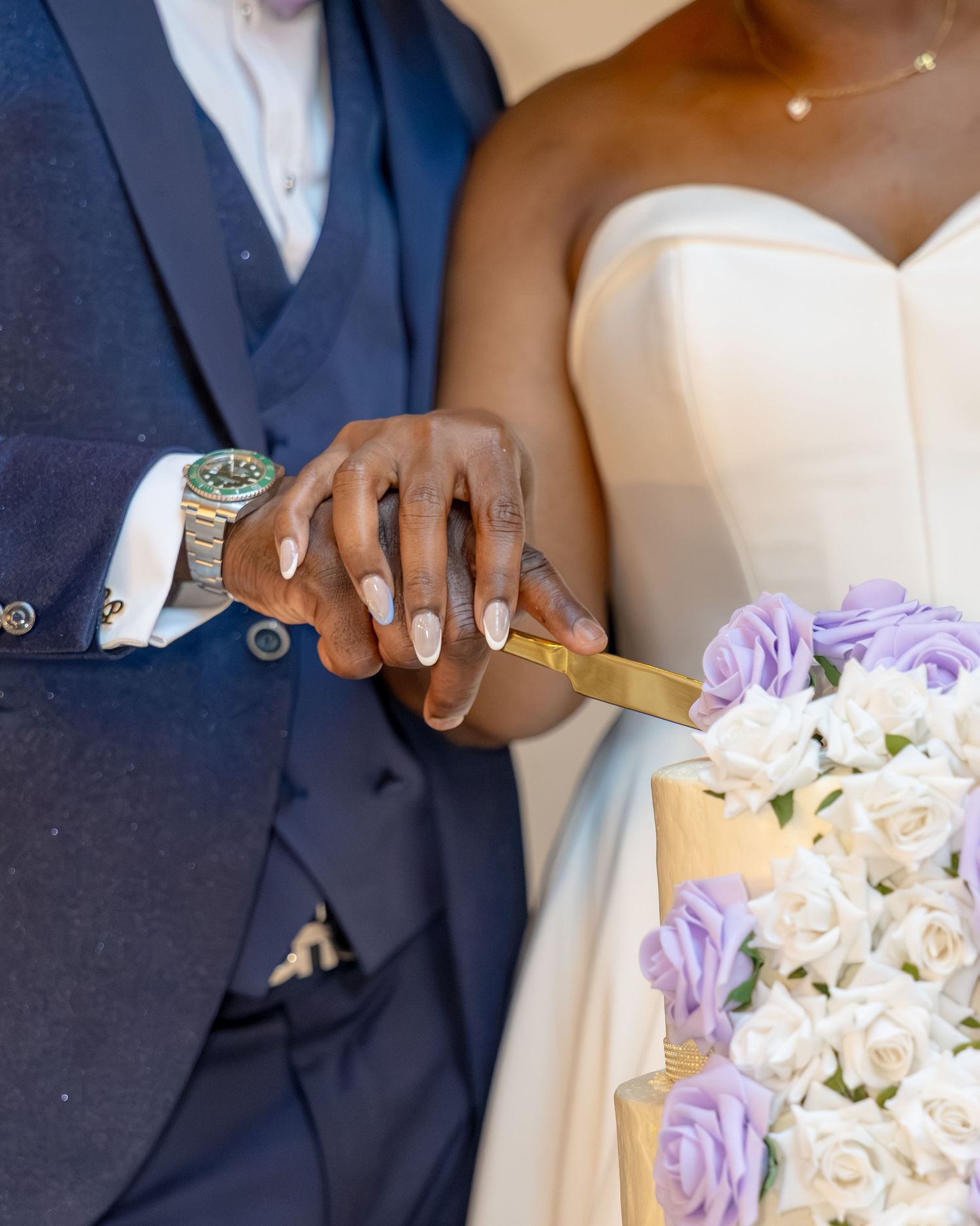A couple cuts their wedding cake together, bride holding white bouquet with purple roses, groom in navy suit with green watch.