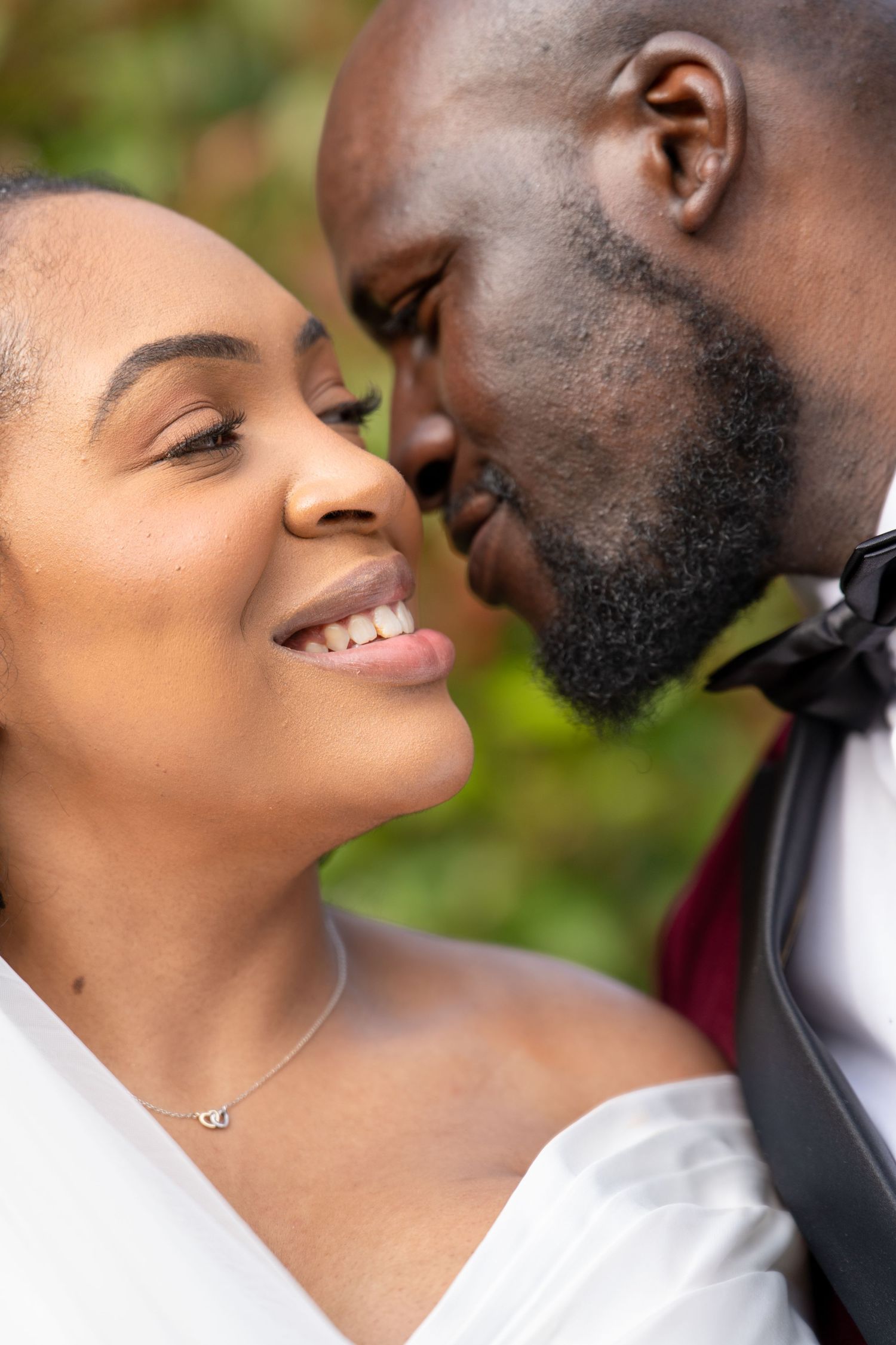 A joyful Black bride smiles as her groom leans in for a kiss on their wedding day outdoors.