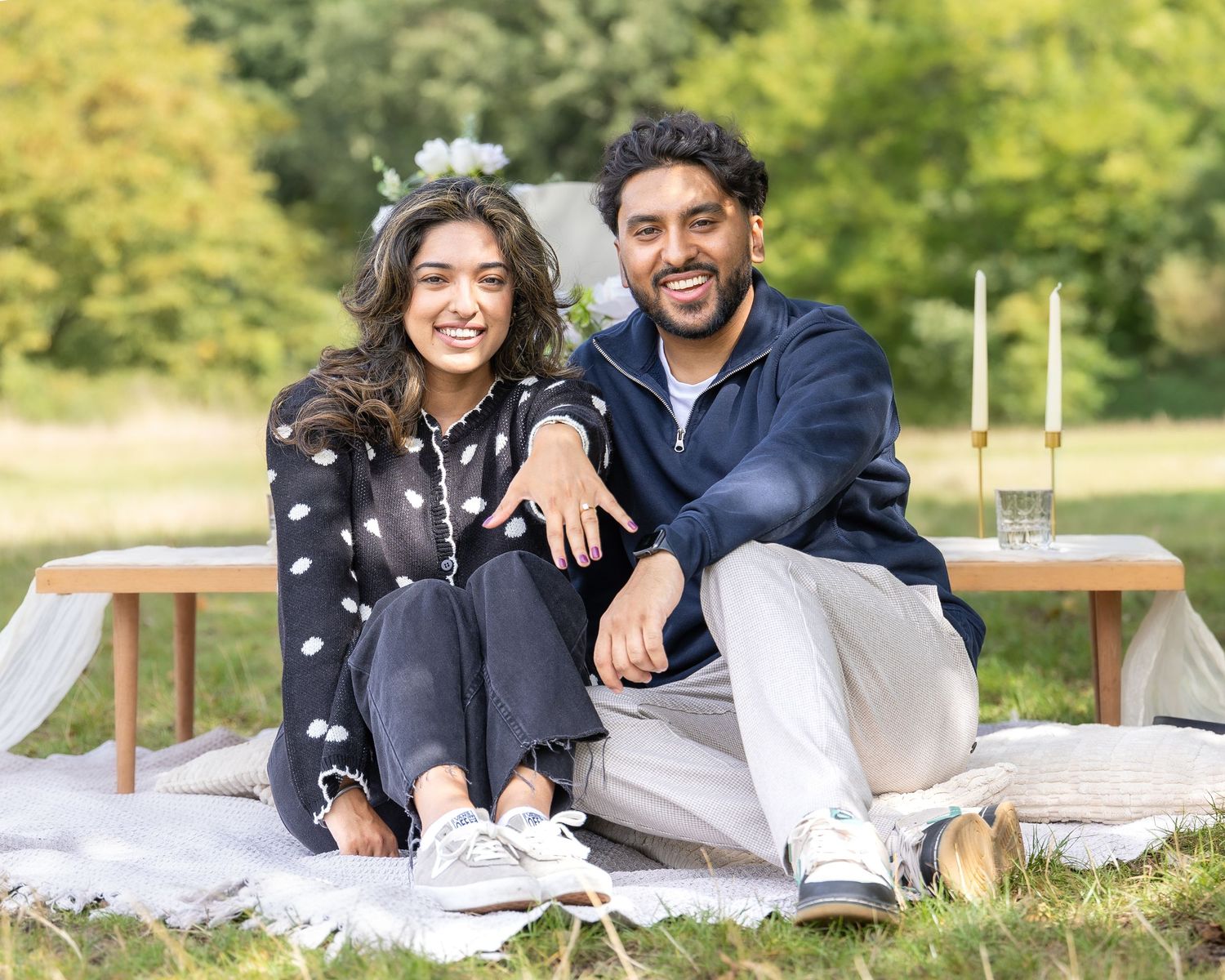 Smiling engaged couple sitting together at outdoor picnic setup with candles in green park.