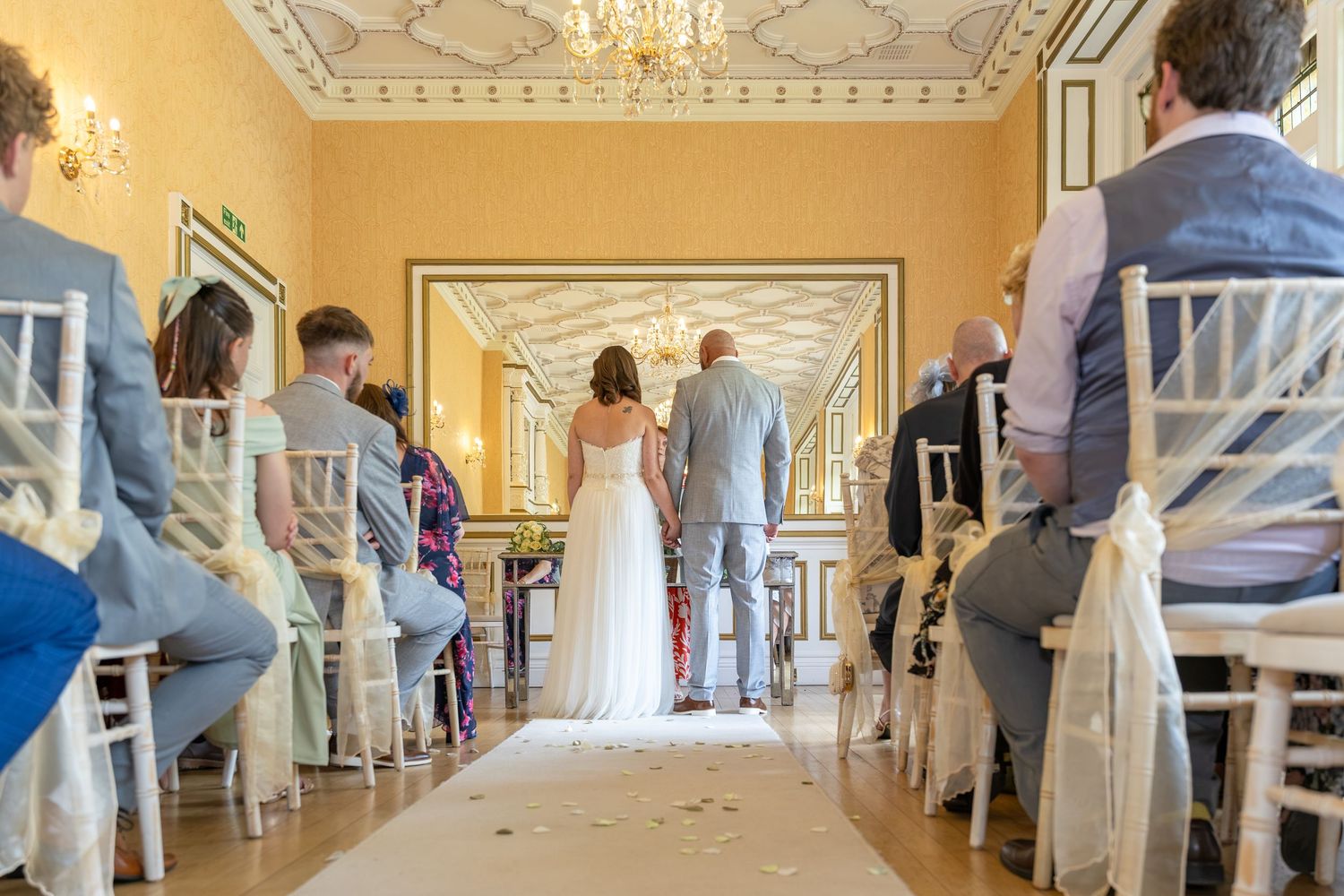 Elegant wedding ceremony in a yellow ornate room with bride and groom at the altar, guests seated on white chairs.