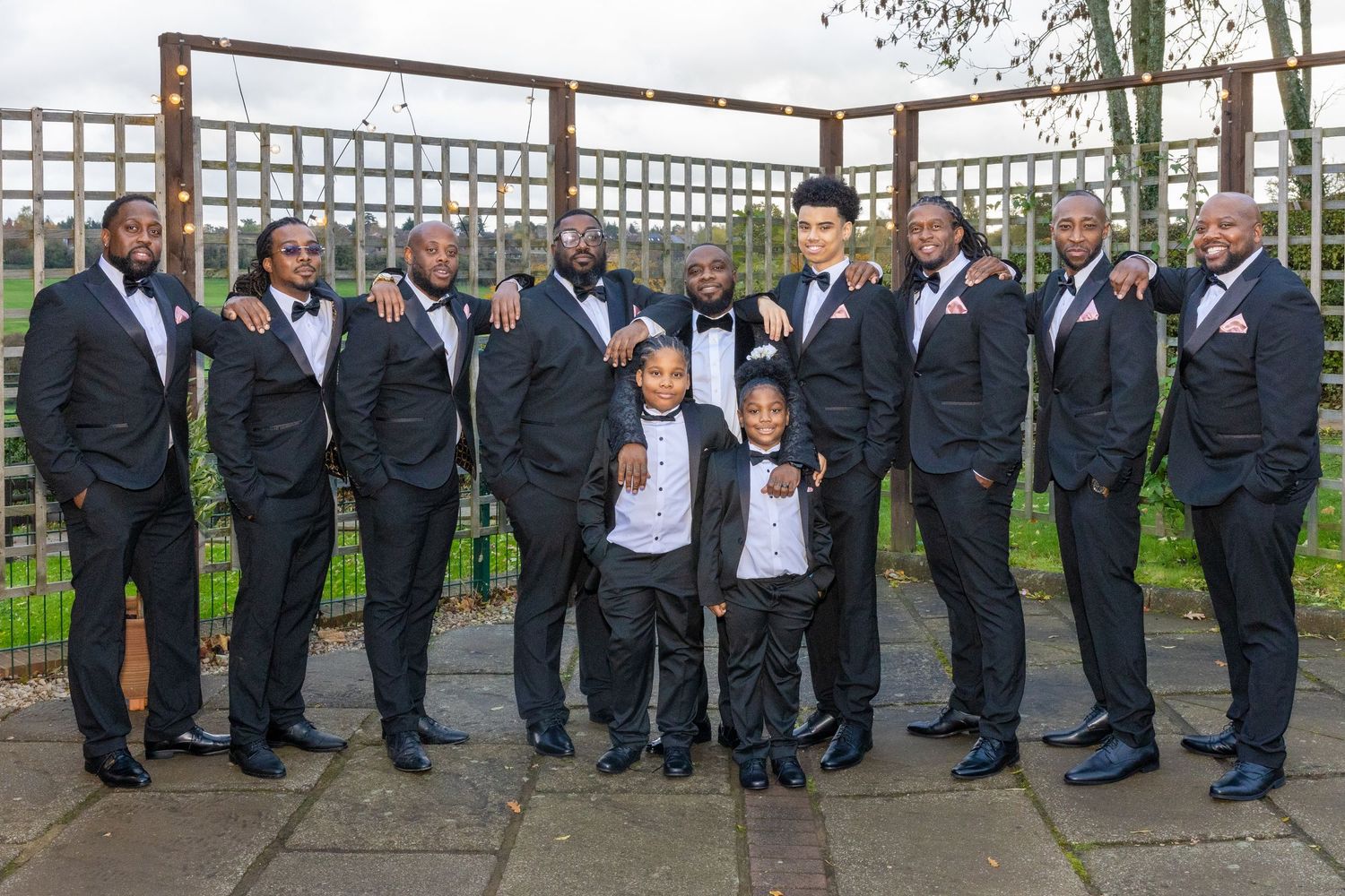 Groom with groomsmen and two young boys in matching black tuxedos pose formally at an outdoor wedding venue.