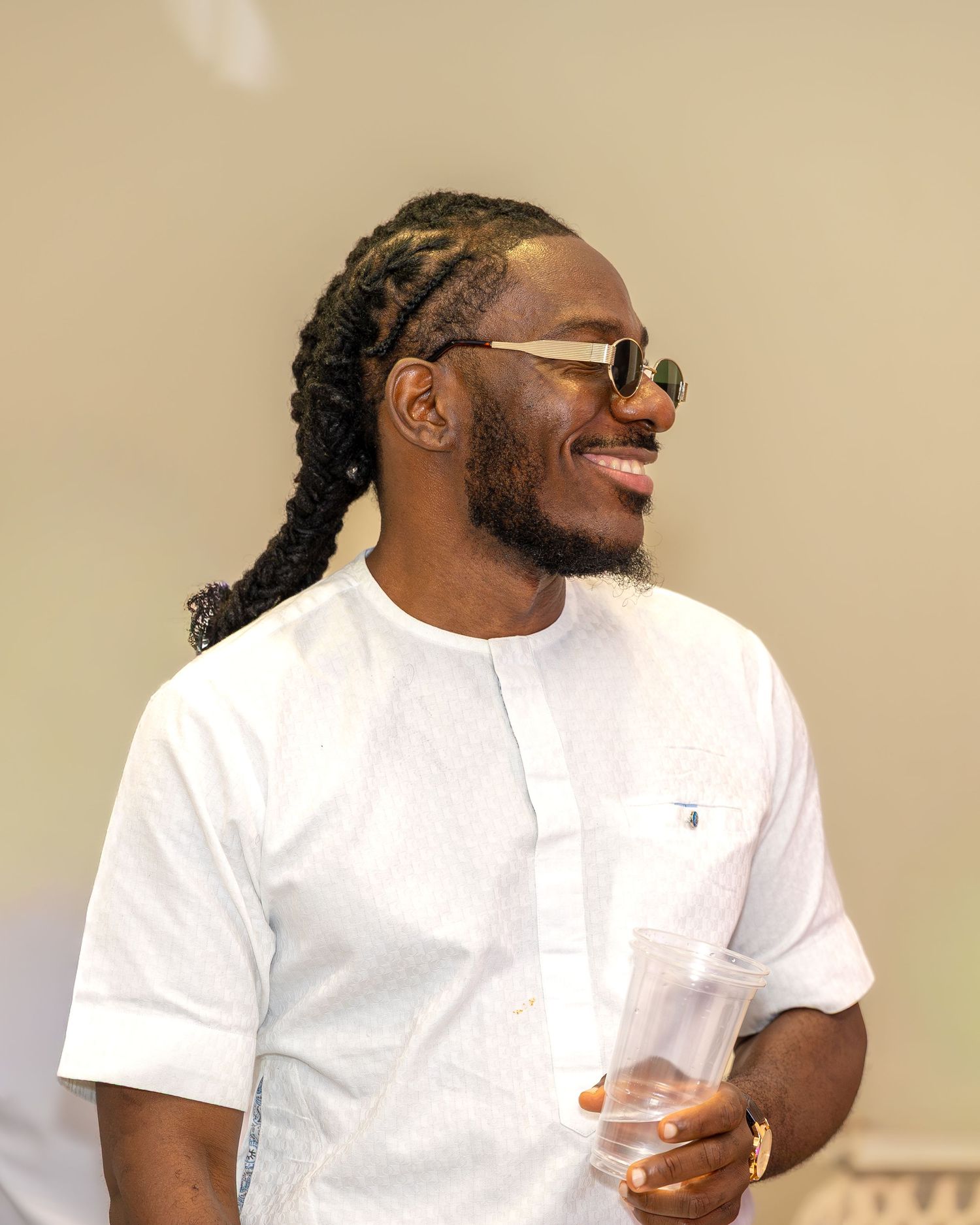 Smiling man with long locs and sunglasses in white Nigerian traditional attire holding drink at wedding celebration.