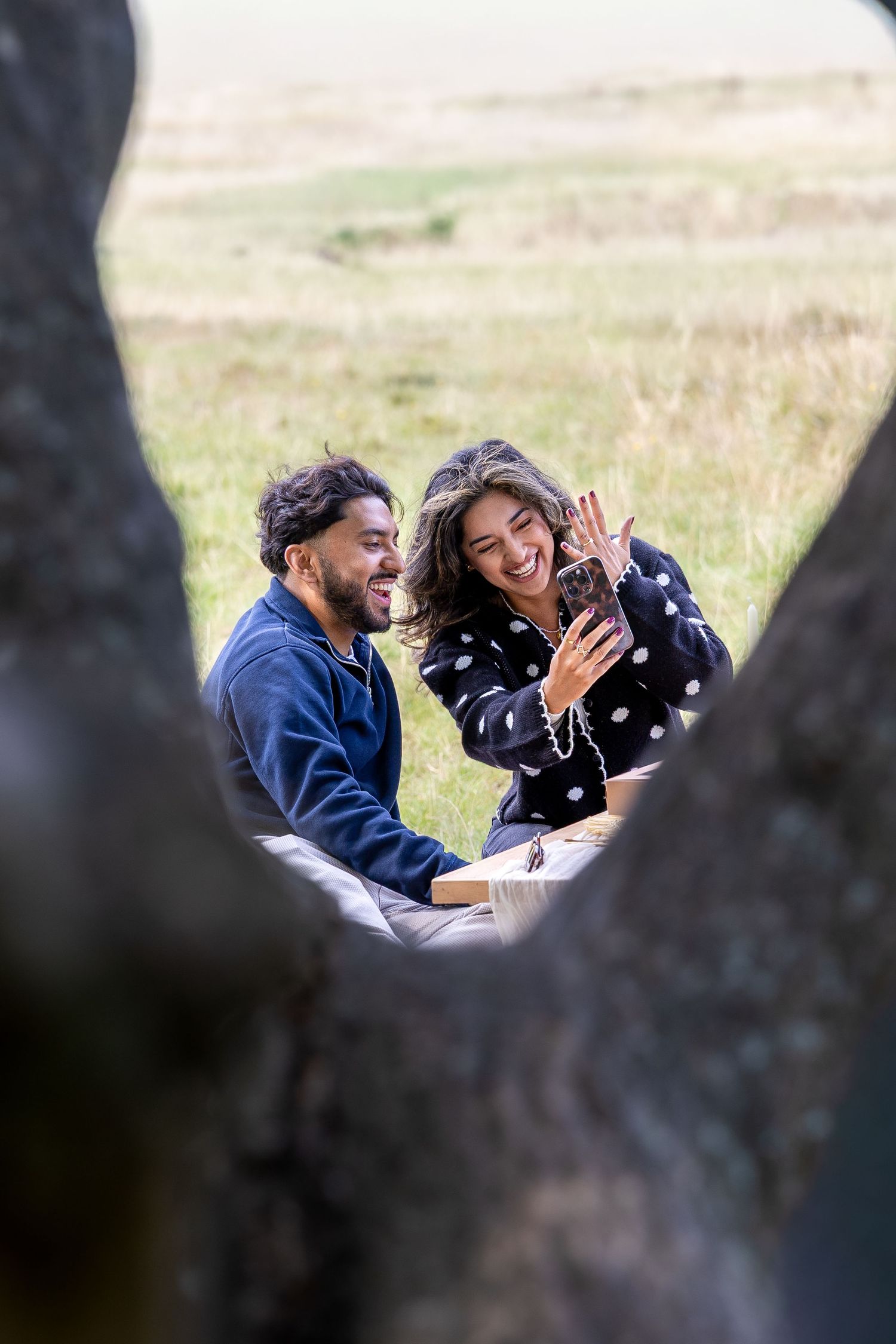Couple laughing during outdoor picnic proposal, woman showing engagement ring while sitting in green field.