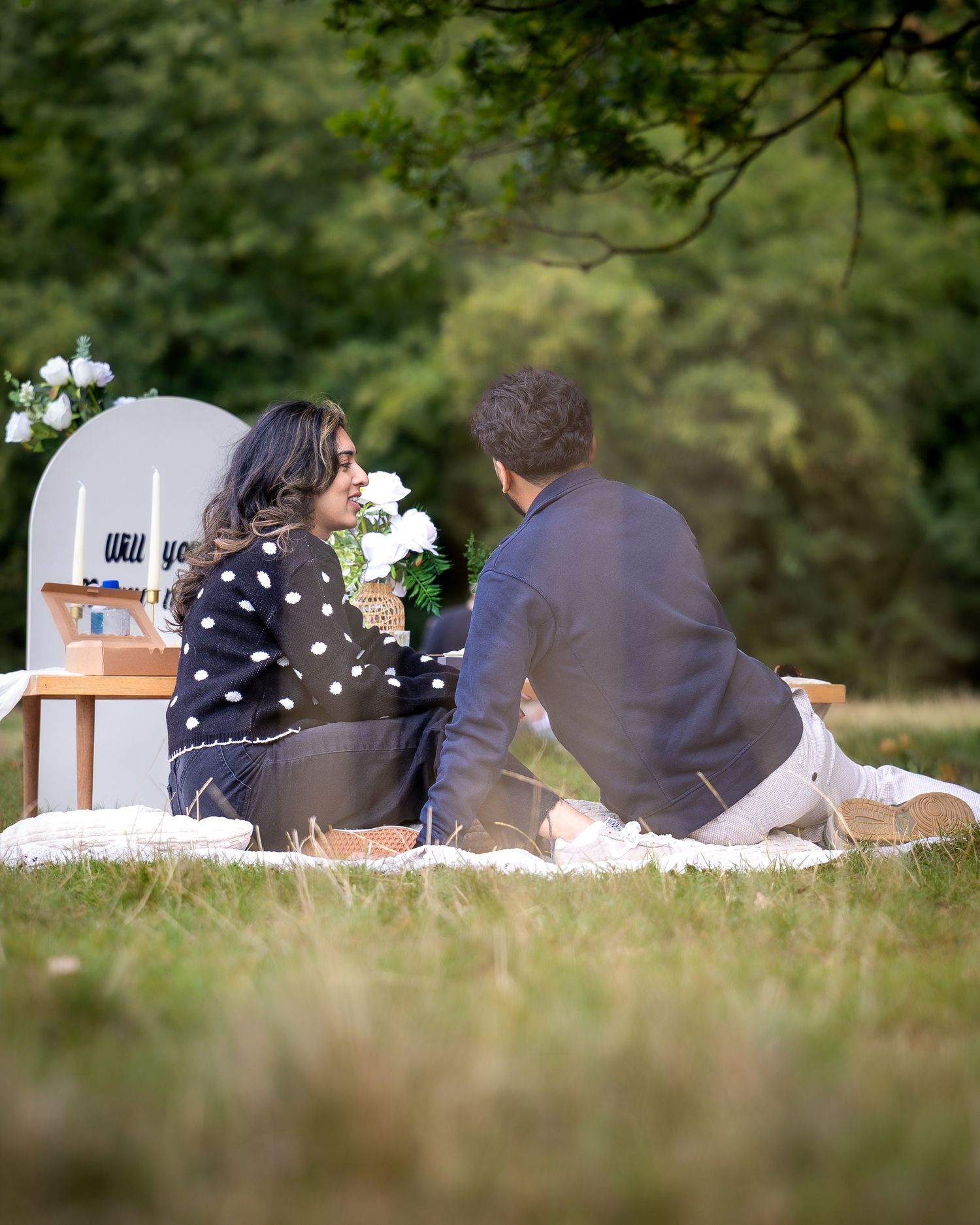 Couple sitting together at outdoor picnic proposal with Will You Marry Me sign and white flowers.