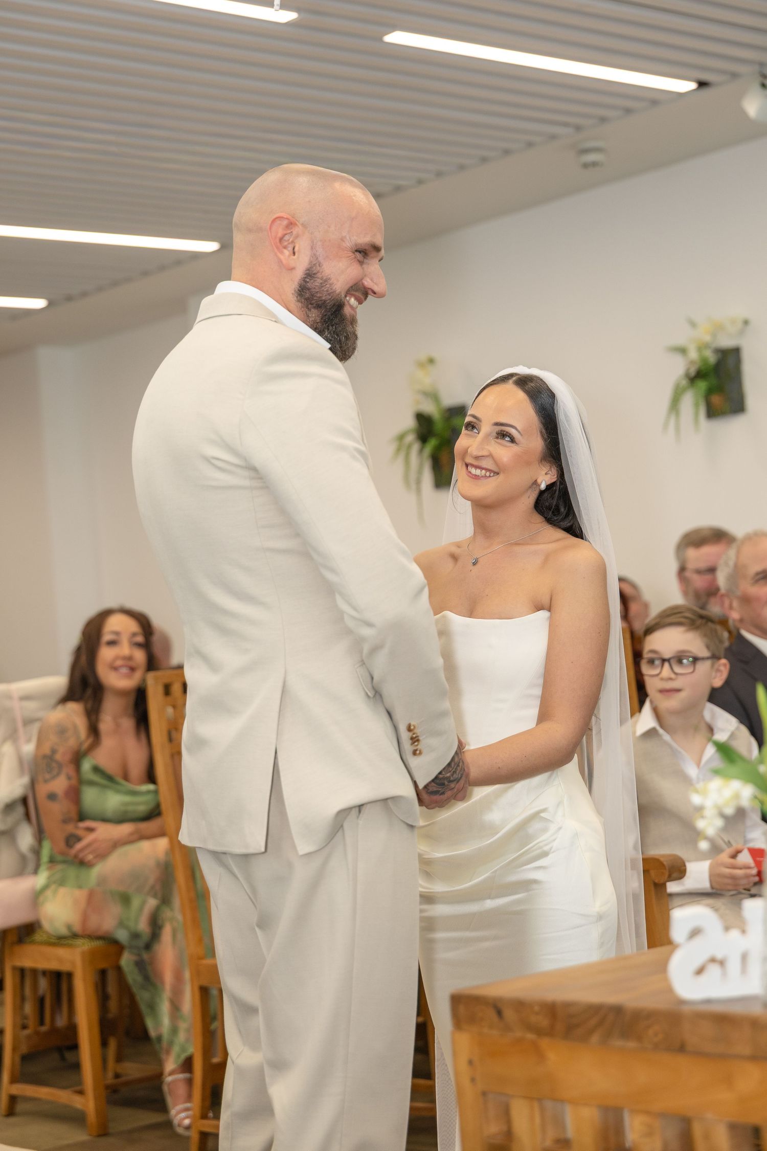 Bride and groom hold hands at altar smiling, with wedding guests seated and laughing in background.