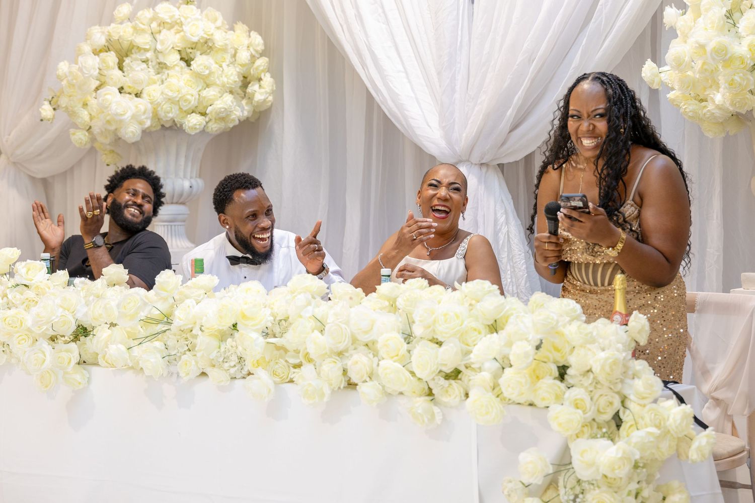 Joyful wedding party laughing at a floral-decorated head table with white roses and elegant draping.