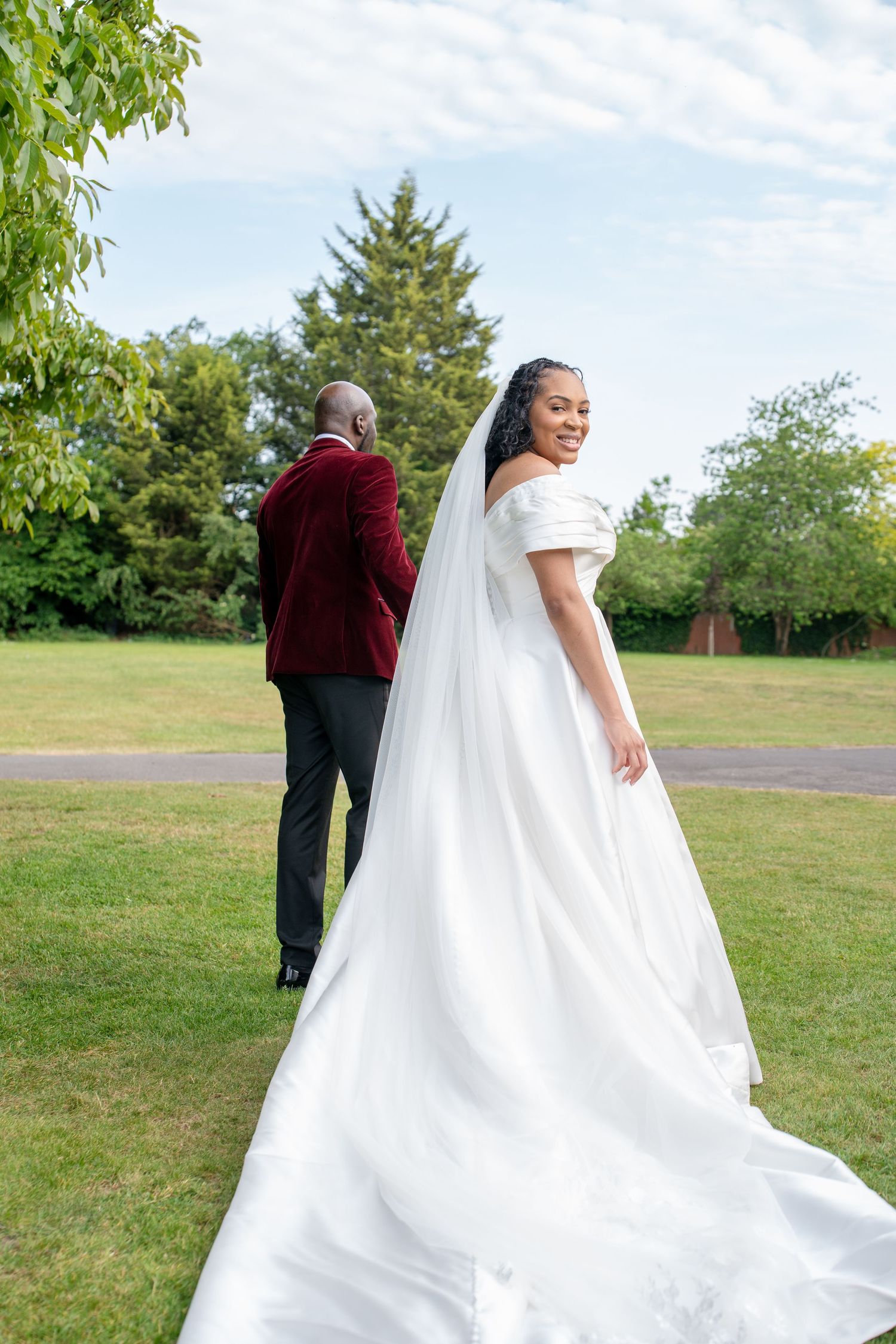 Bride in white off-shoulder gown with long veil smiles back at camera while groom in burgundy jacket stands in lush garden.