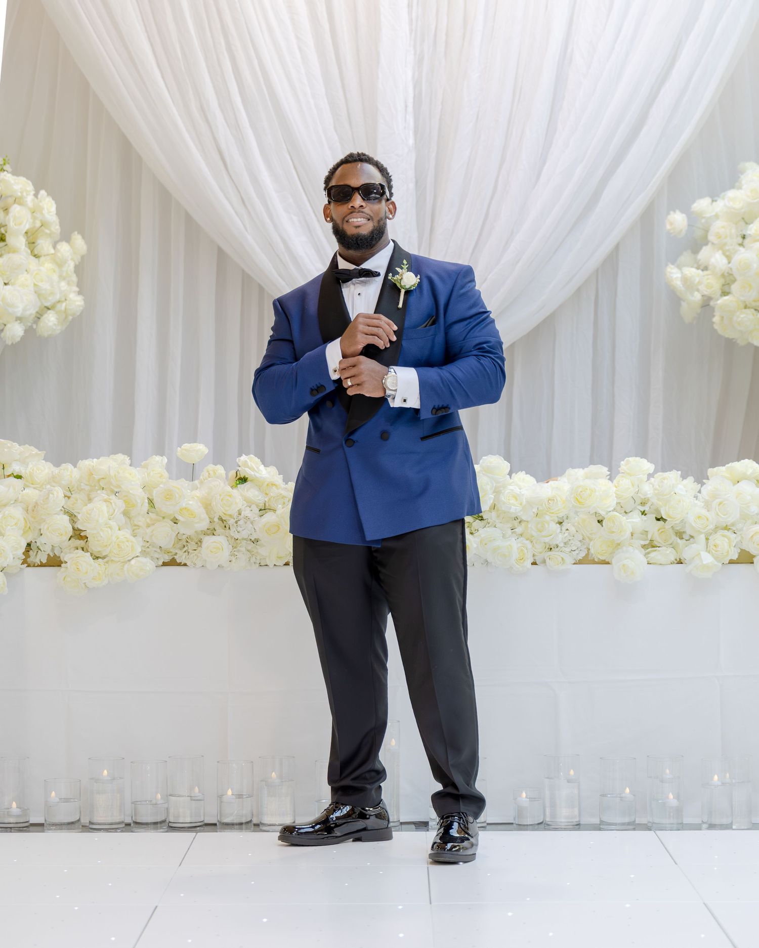 Man in blue tuxedo jacket with black bow tie and sunglasses posing at elegant wedding venue with white floral decor.