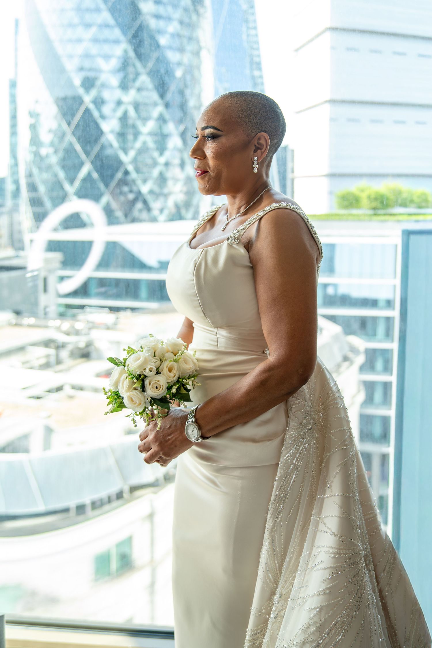 Elegant bald bride in ivory gown holding white bouquet, standing by window with London cityscape background.