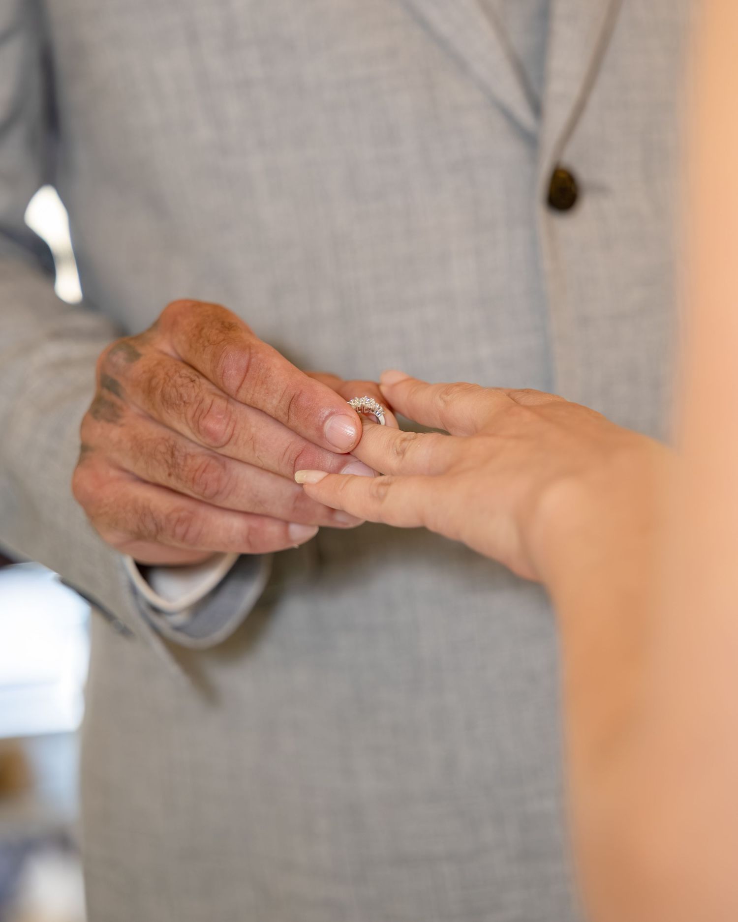 Groom in gray suit placing a diamond wedding ring on bride's finger during wedding ceremony.