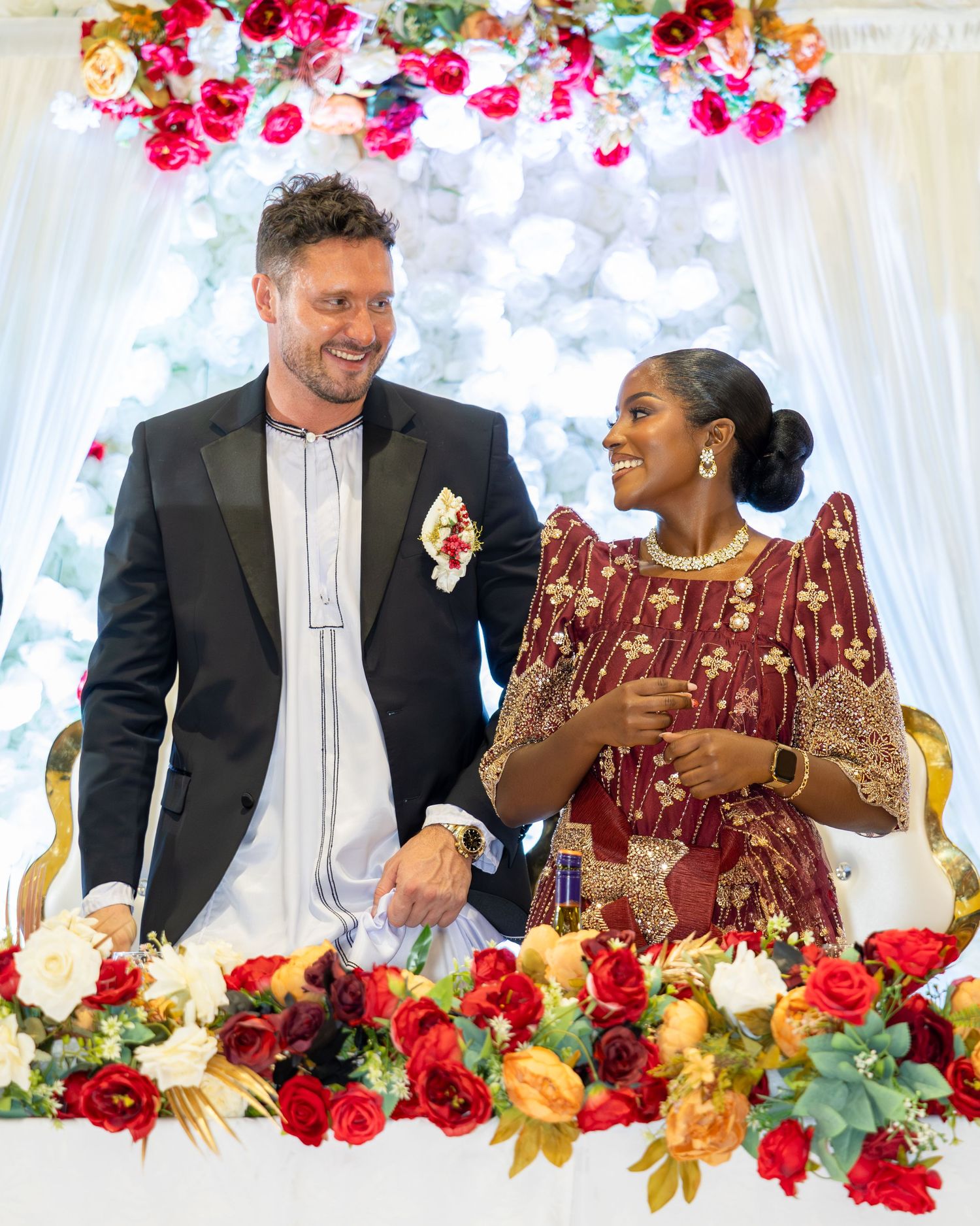 Interracial couple smiling at each other at wedding altar surrounded by red and white floral arrangements.