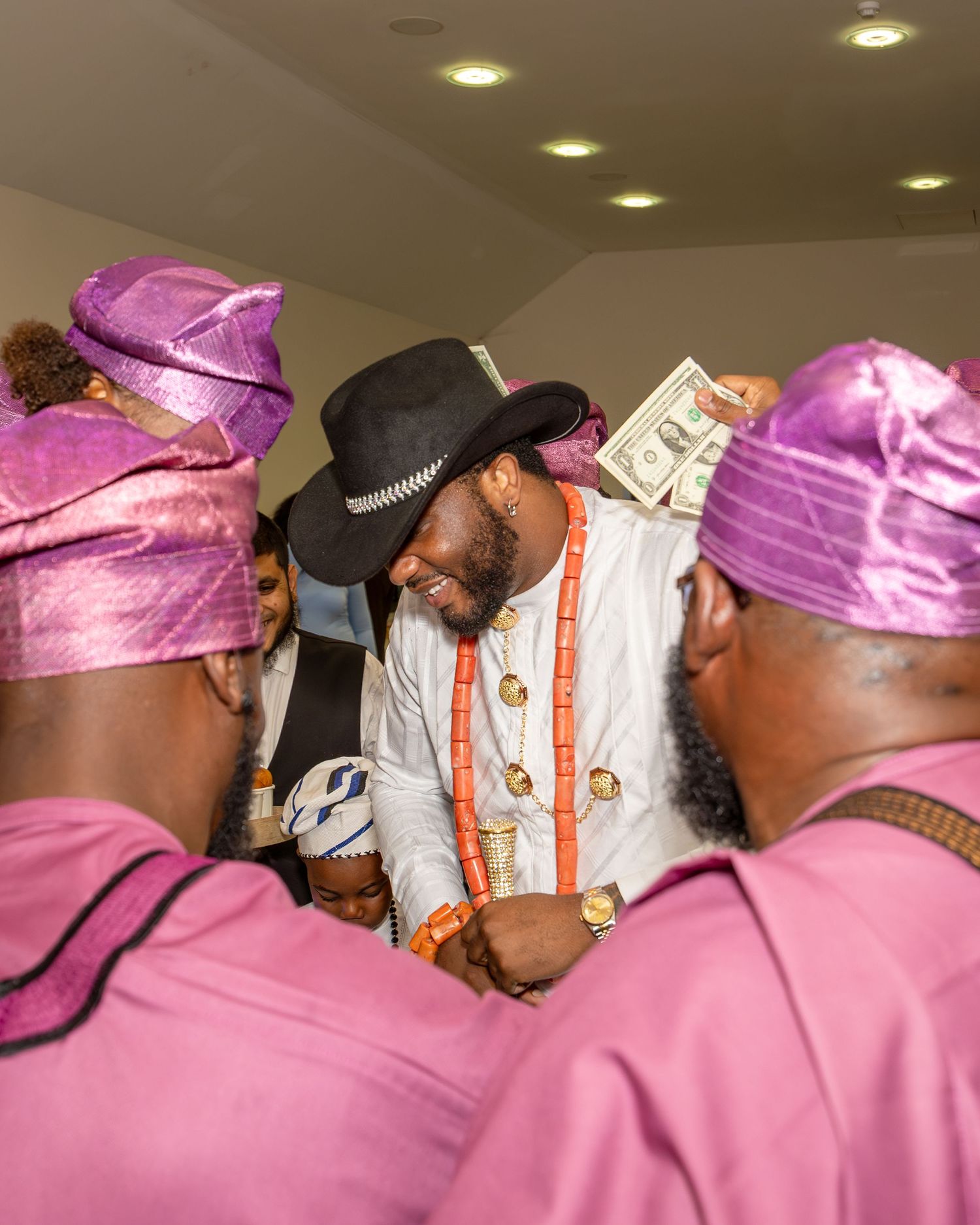 Groom in black cowboy hat and coral beads receives money spray from guests in pink aso-oke at Nigerian wedding.