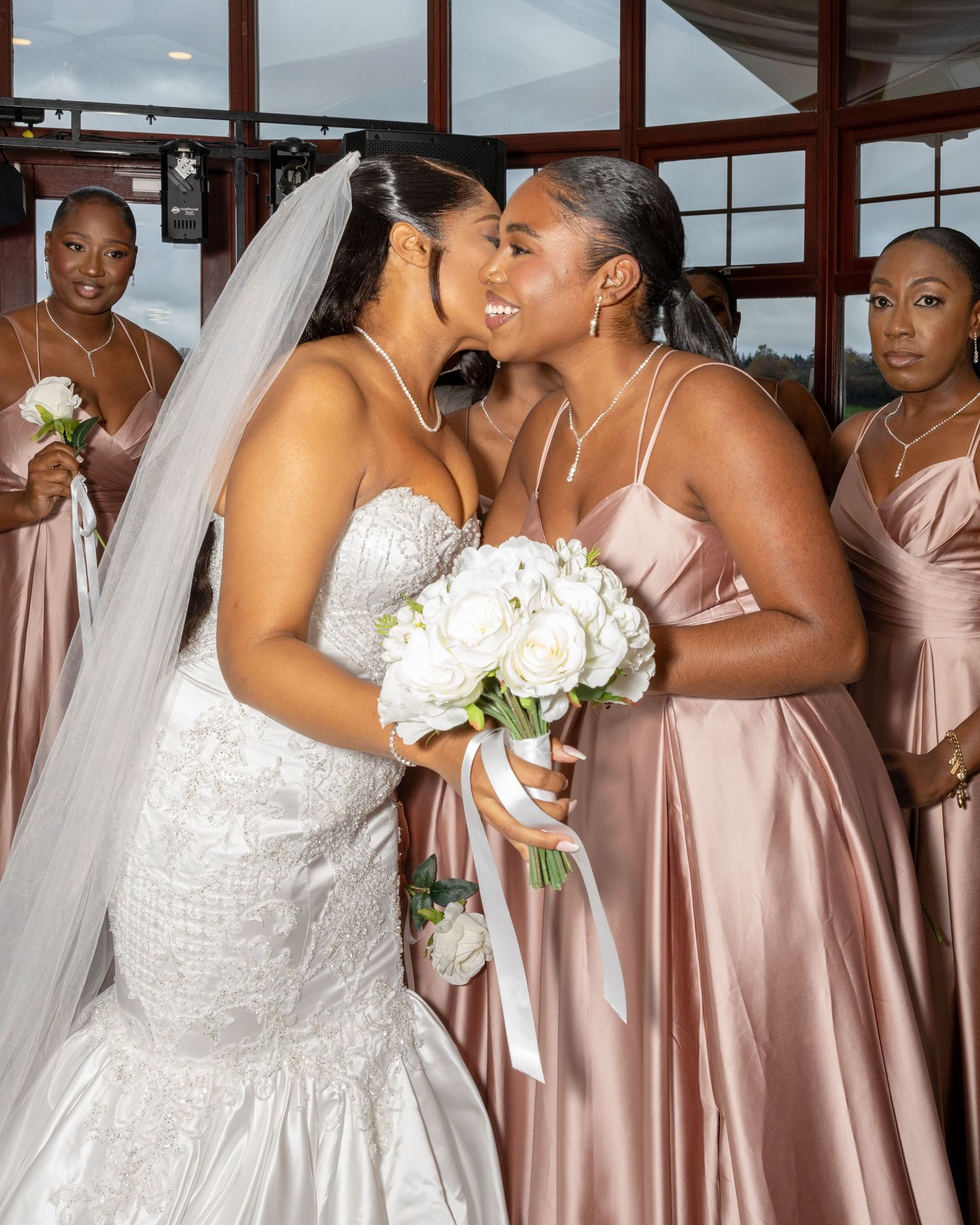 Bride in white lace gown embracing bridesmaid in champagne dress at wedding reception.