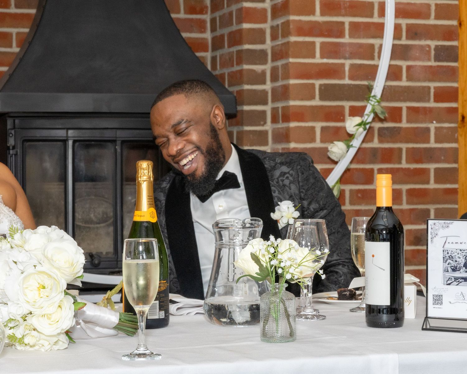 Groom in patterned tuxedo laughs joyfully at the wedding reception head table with champagne nearby.