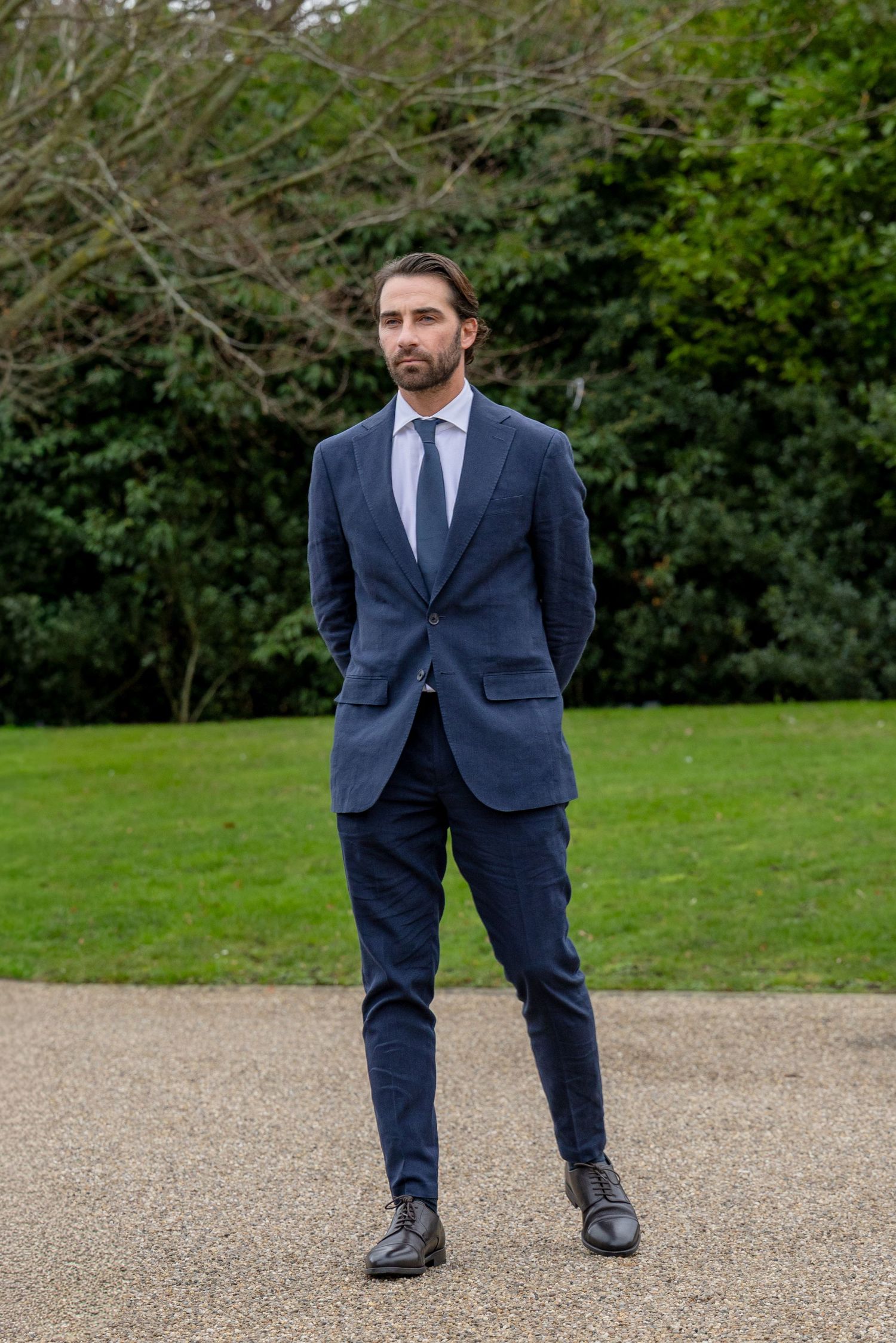 Stylish man in a navy blue suit standing on a gravel path in a lush green garden.