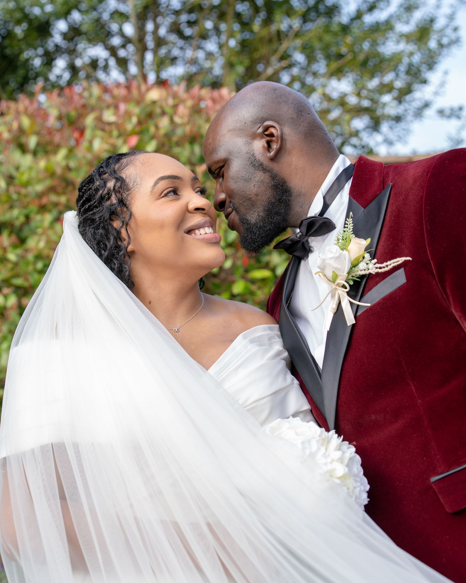 A joyful bride in a white gown and veil shares a romantic moment with her groom in a burgundy velvet tuxedo outdoors.