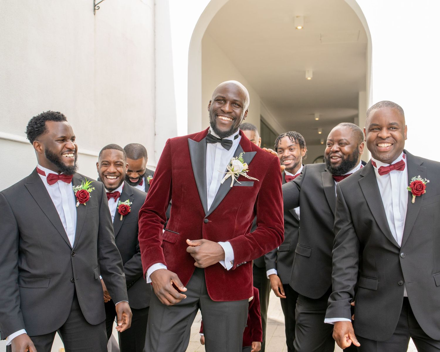 Groom in burgundy velvet tuxedo jacket poses with groomsmen in gray suits and red bow ties at wedding.
