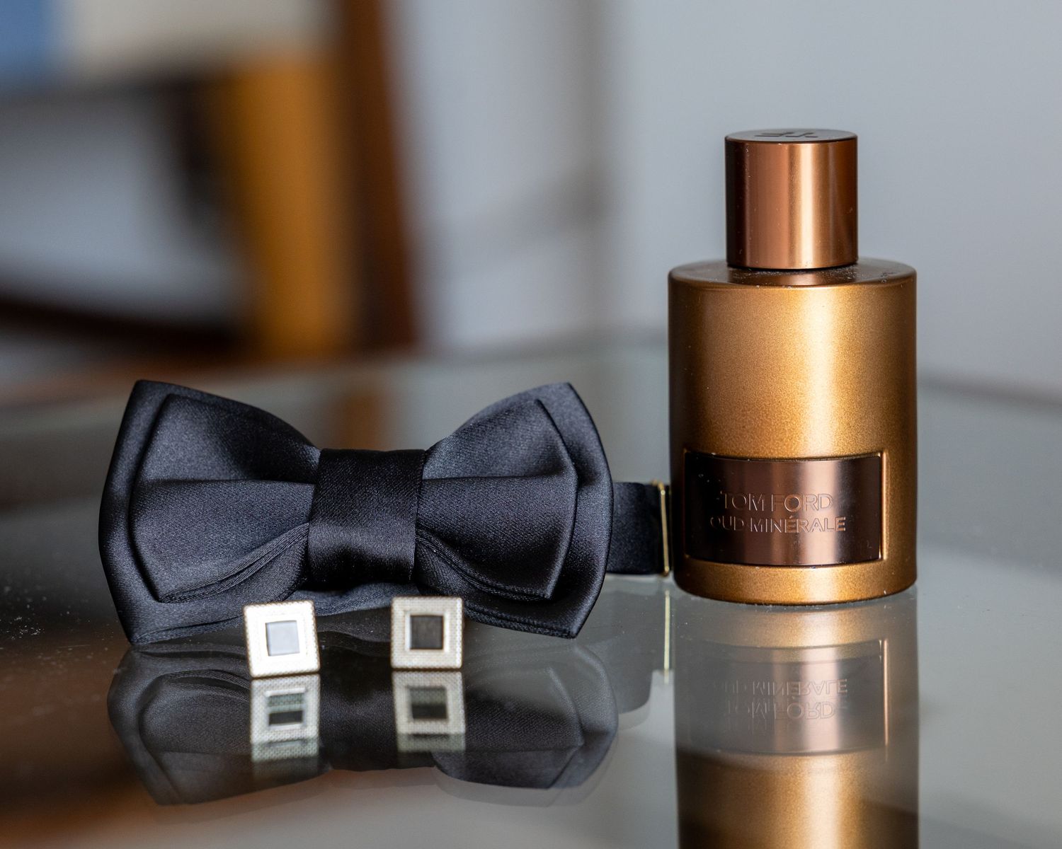 Black satin bow tie, silver cufflinks, and bronze cologne bottle arranged elegantly on a reflective surface.