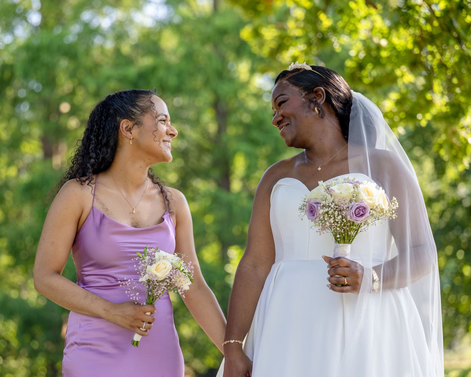 Bride in white gown and bridesmaid in purple dress holding bouquets, smiling outdoors with green trees.