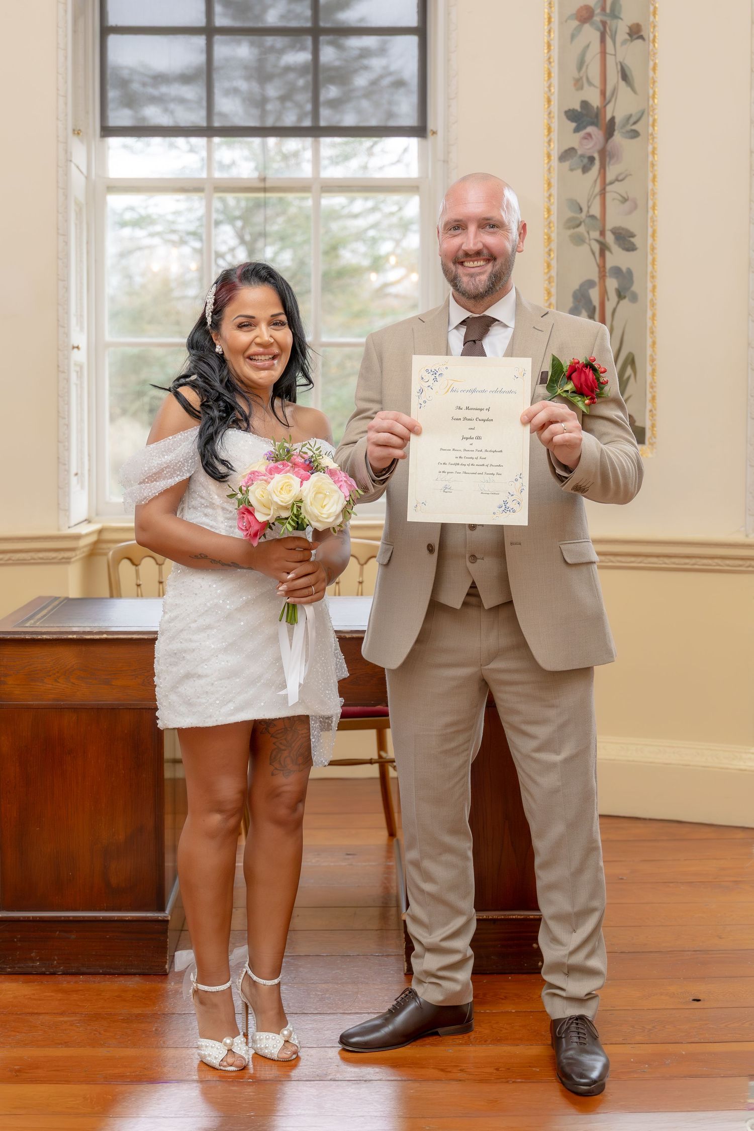 Smiling newlyweds display marriage certificate, bride holds pink bouquet in white mini dress.