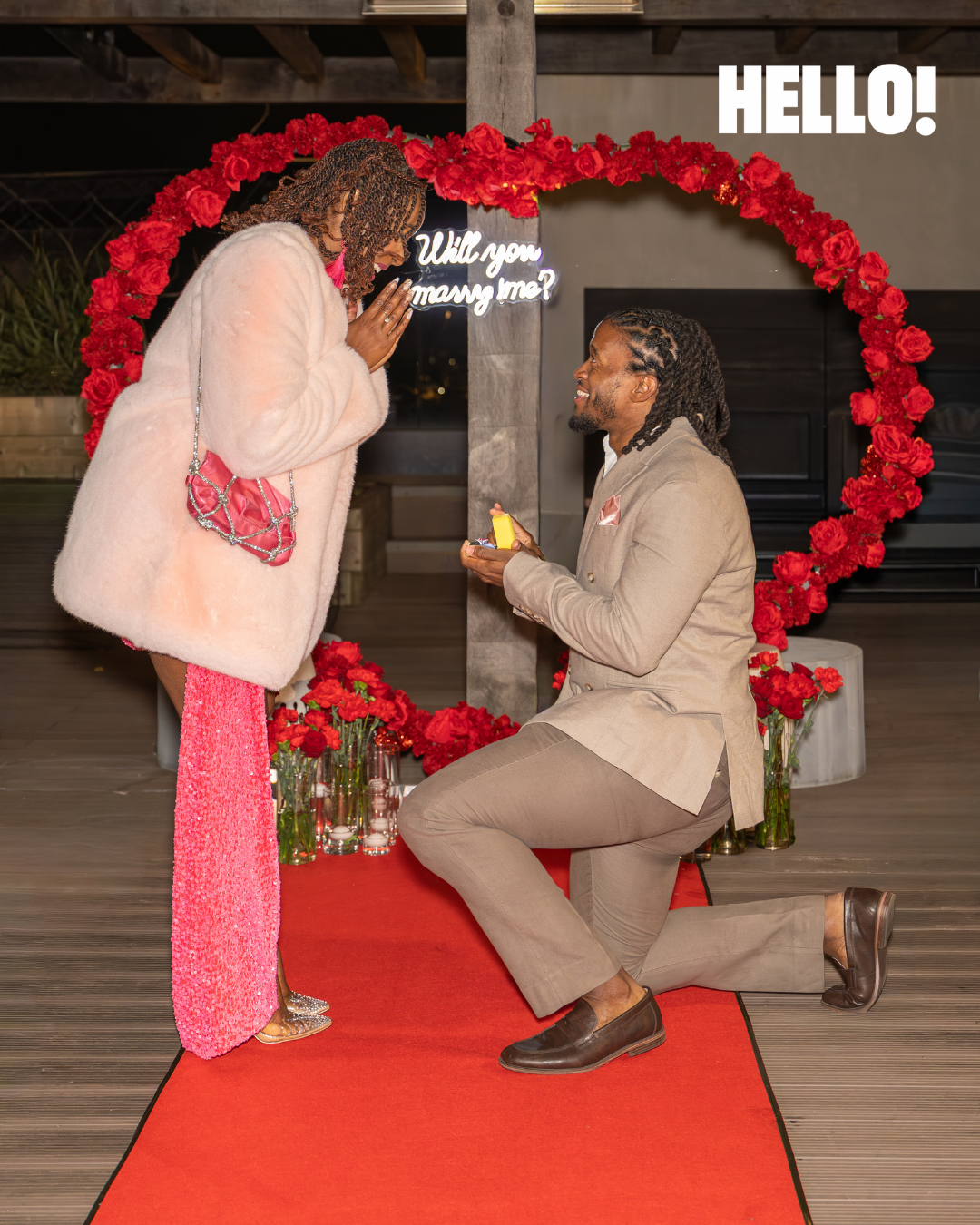 Man proposes marriage to woman in front of red rose heart arch with Will You Marry Me neon sign.