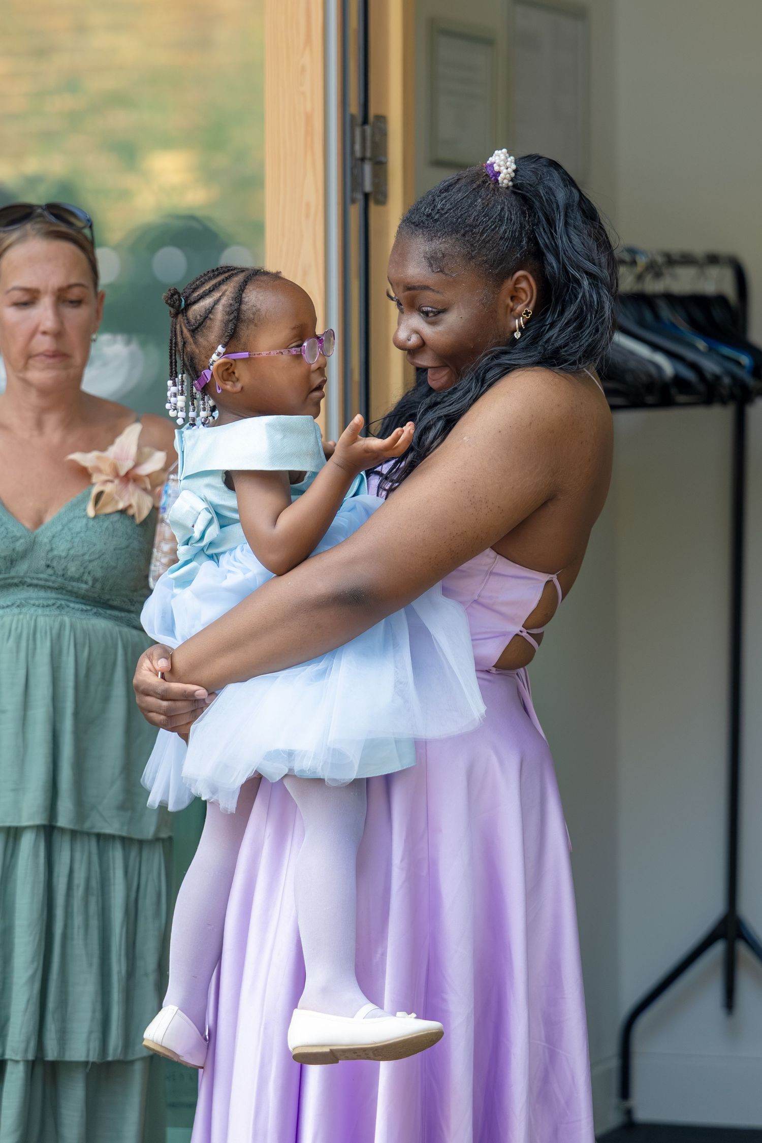 Woman in lavender gown holds young girl in blue tulle dress at an outdoor wedding celebration.