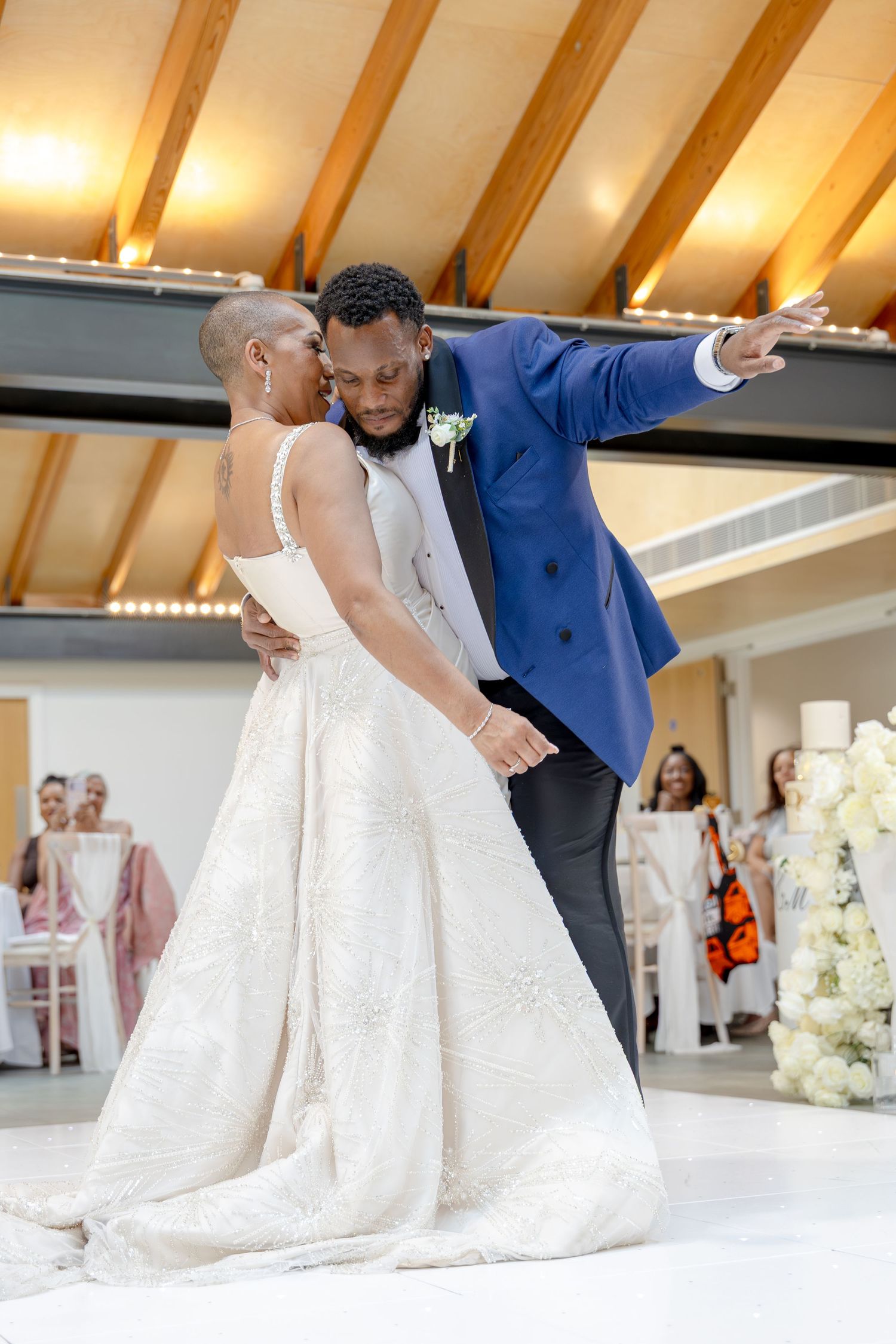 Bride and groom share first dance at wedding reception, groom in blue suit pointing while holding bride in white gown.