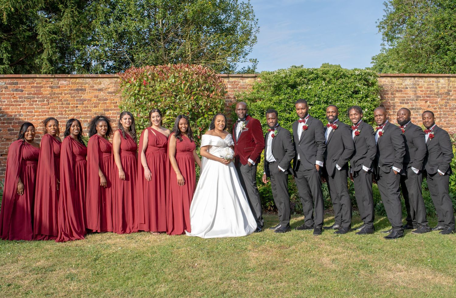 Wedding party poses outdoors, bride in white gown flanked by bridesmaids in burgundy dresses and groomsmen in gray suits.