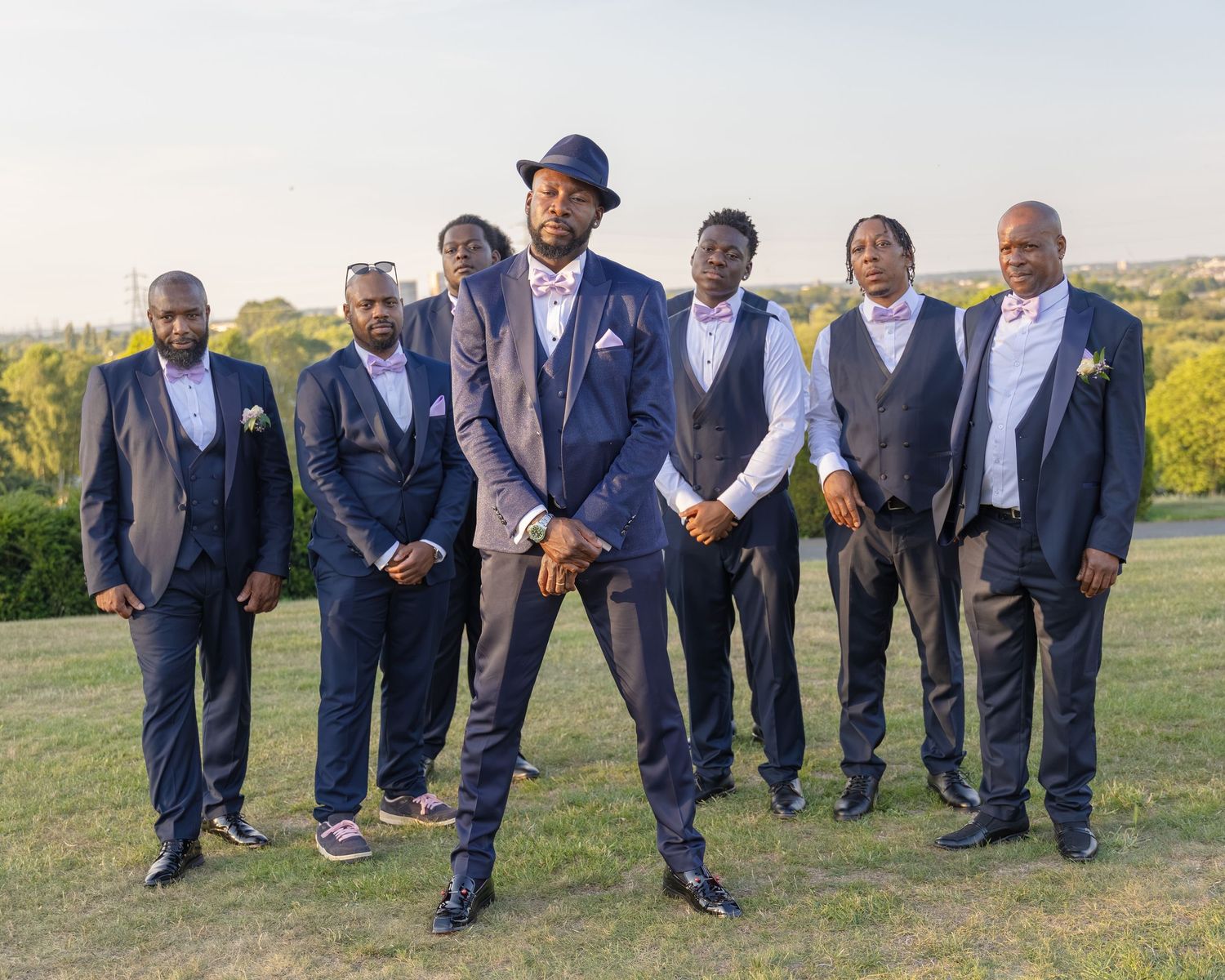 A groom in a navy suit poses confidently with six groomsmen in matching dark suits on a green hillside.