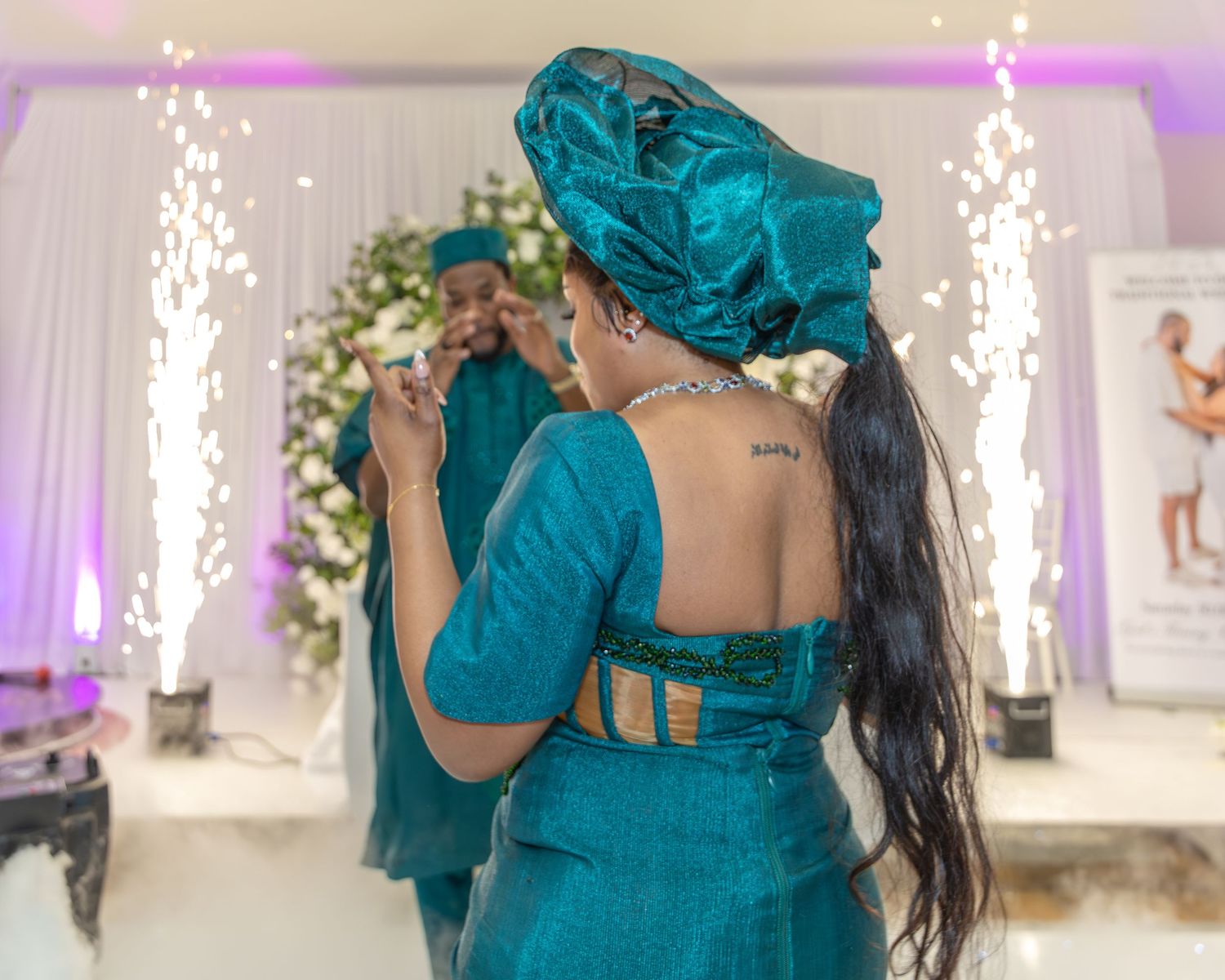 A woman in a teal Nigerian traditional outfit faces a mirror with sparklers glowing in the background at a reception.