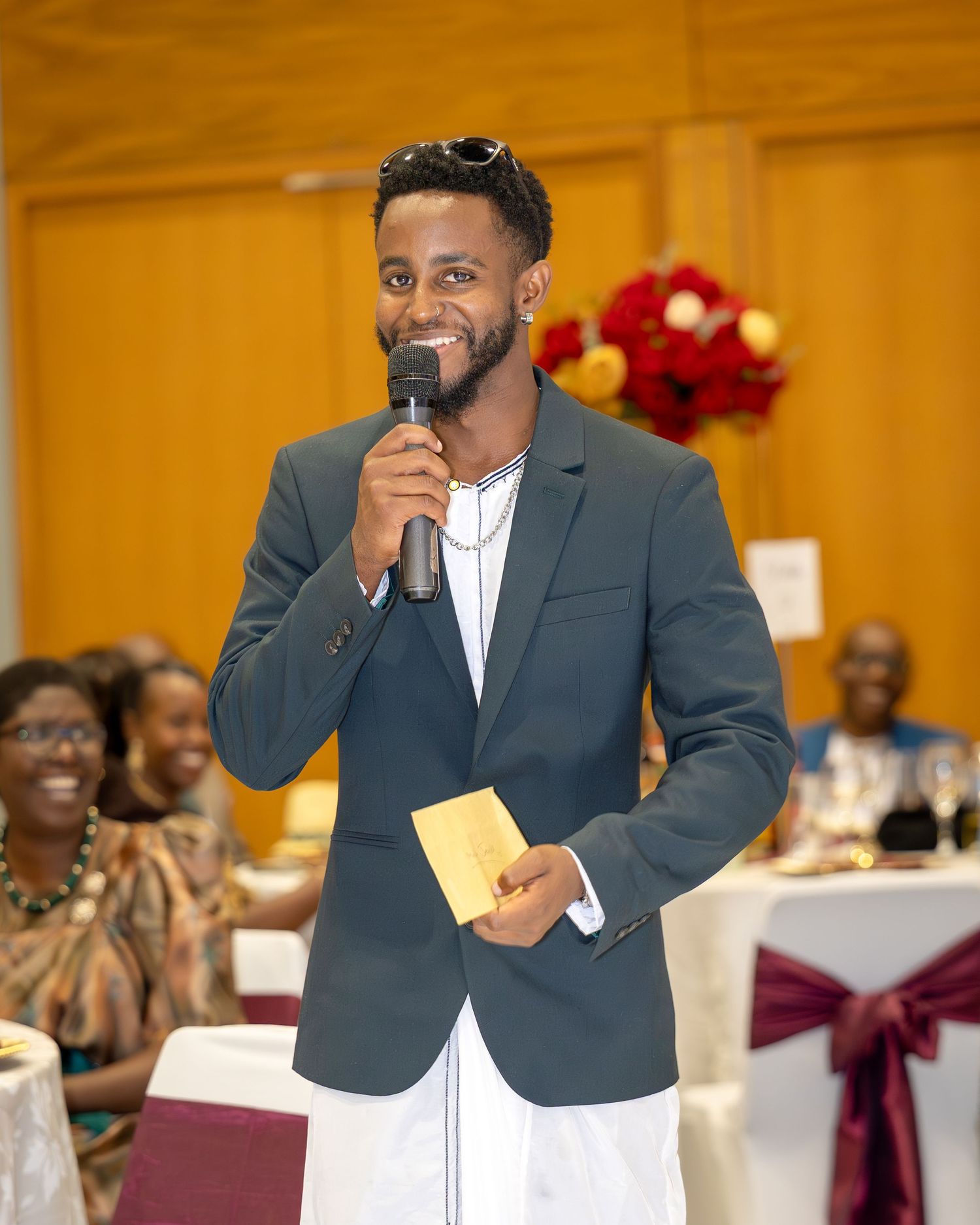 Smiling man in gray blazer speaking into microphone at a wedding reception holding a card.