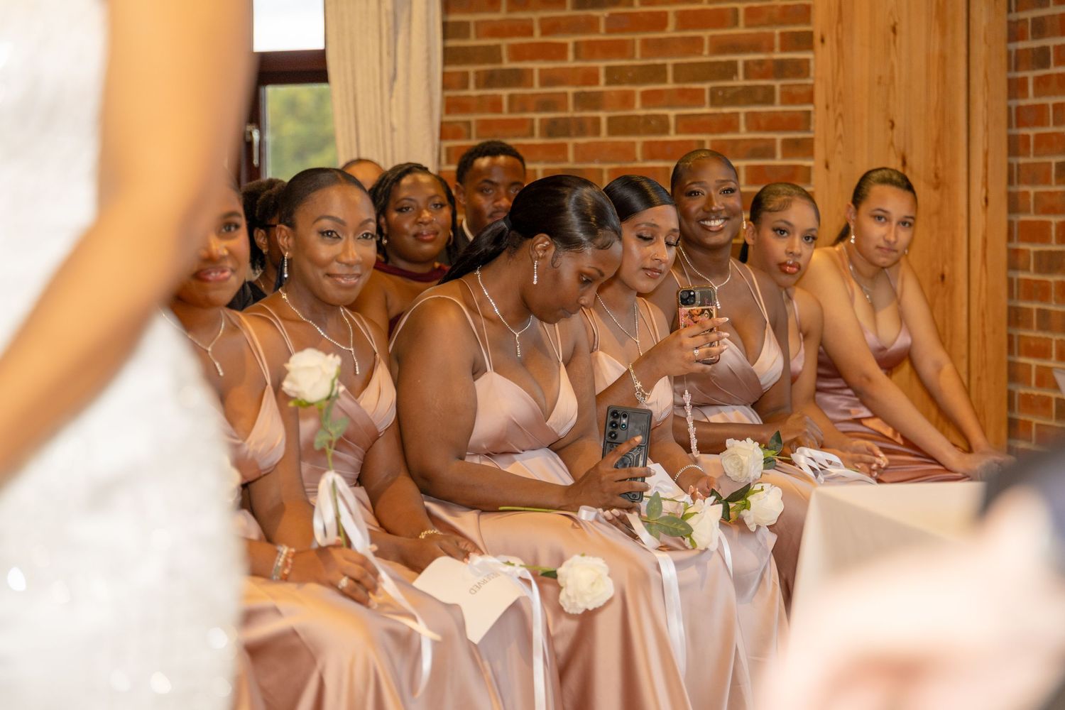 Bridesmaids in matching rose gold satin dresses smile and watch attentively during a wedding ceremony while seated together.