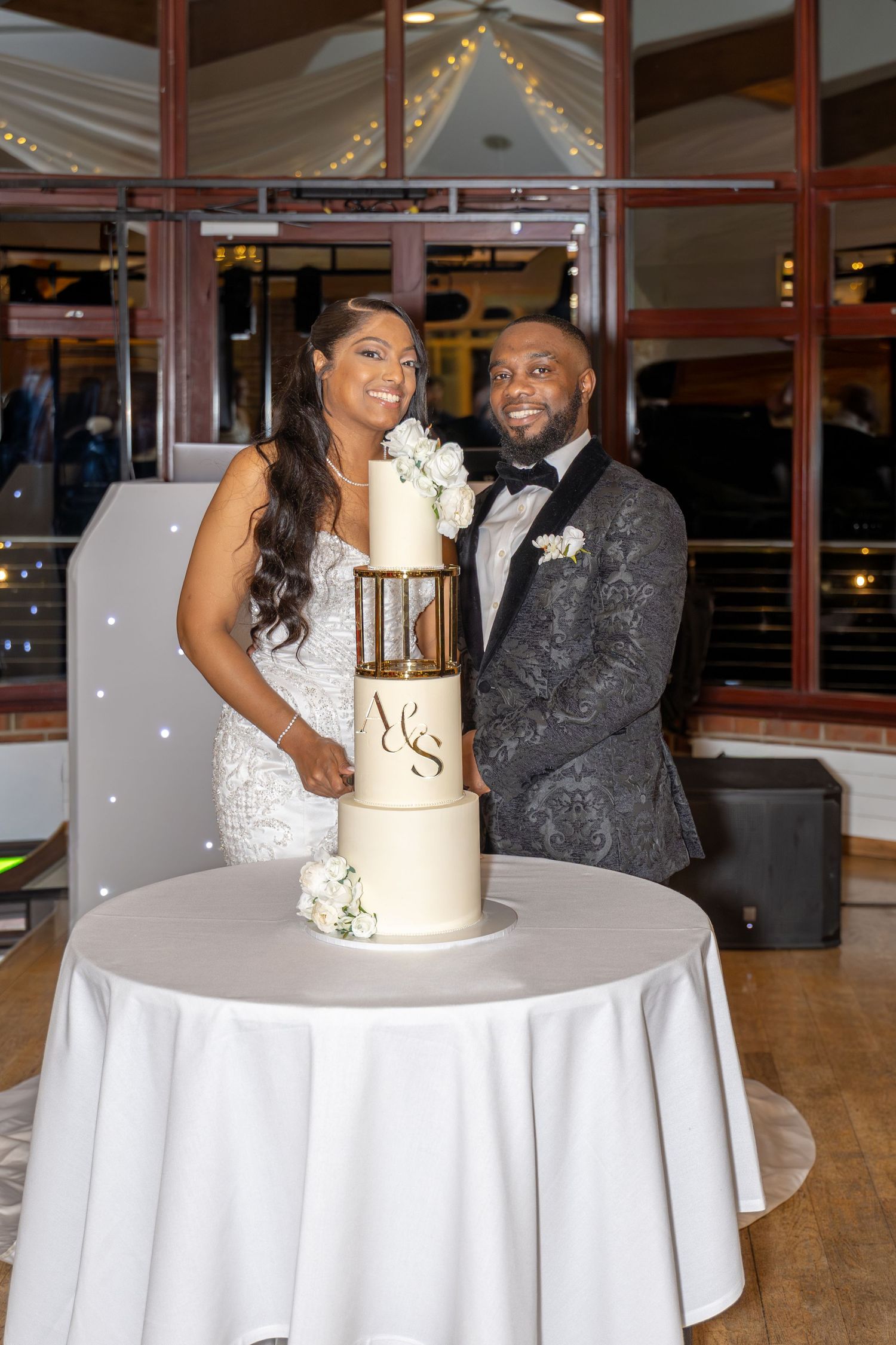 Bride and groom pose beside their elegant three-tier wedding cake with gold monogram initials A and S.