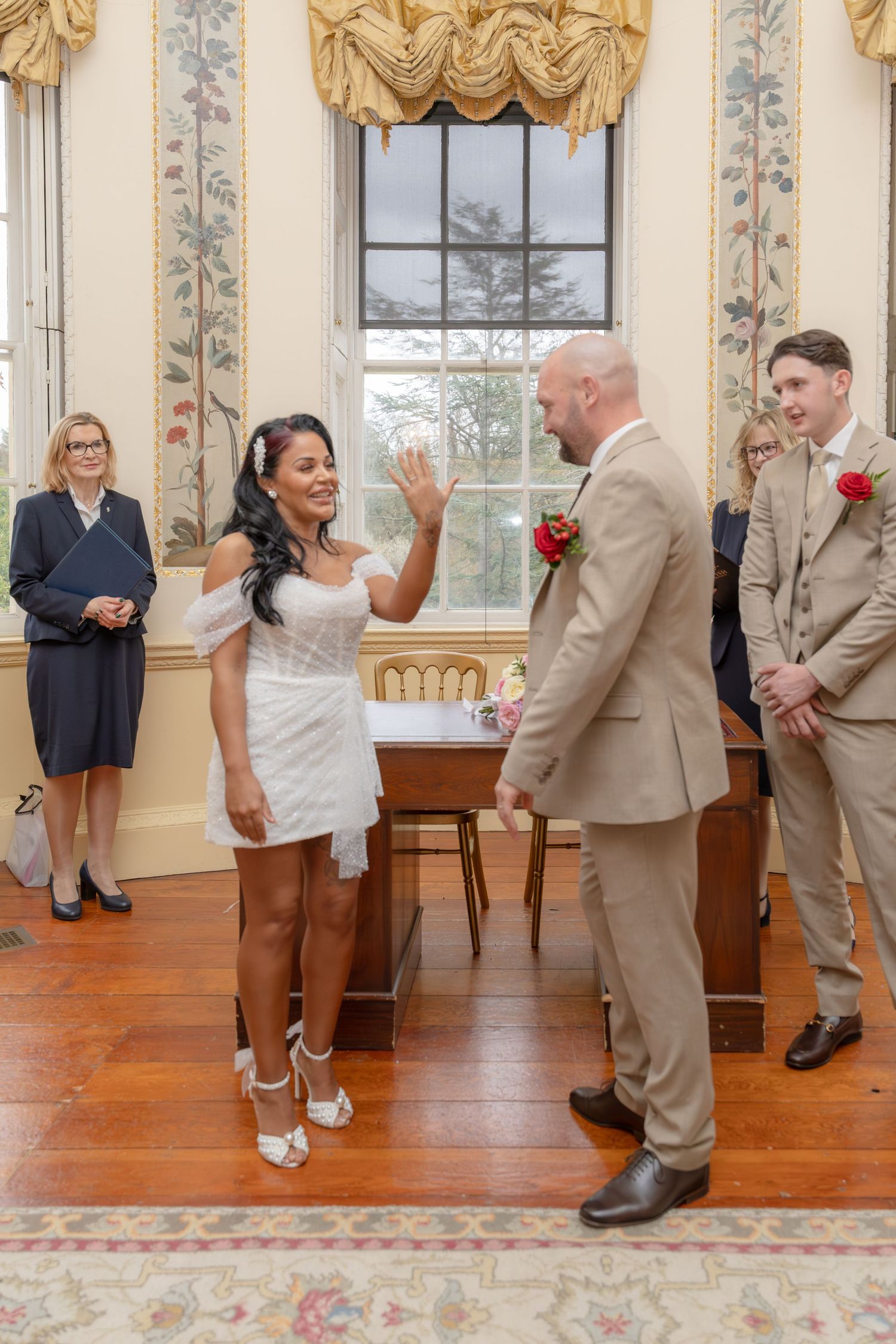 Bride joyfully raising hand after wedding ceremony, groom in beige suit smiling, officiant watching nearby.